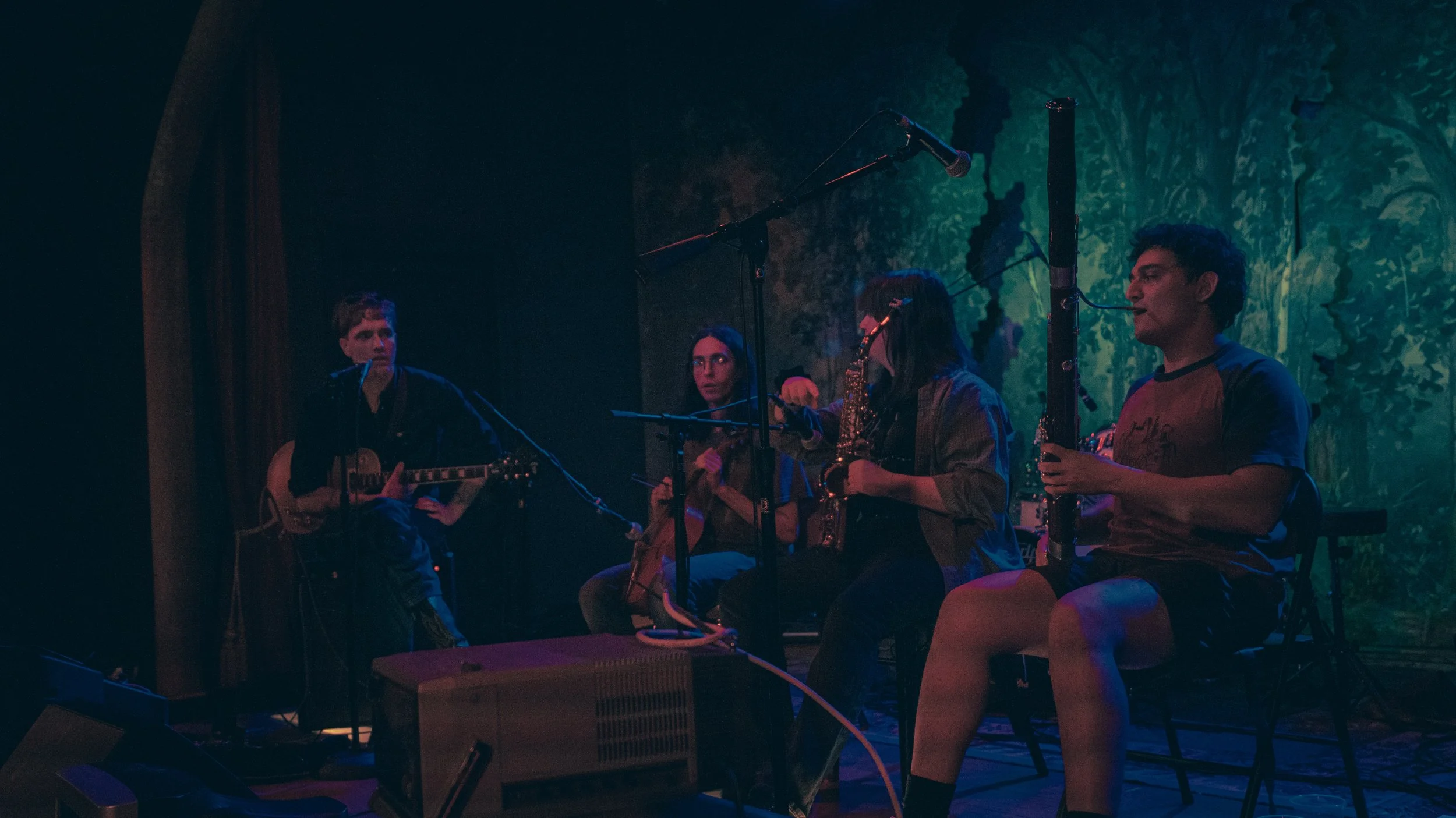 Four musicians performing on stage in a dimly lit, forest-themed setting. One person is playing a guitar, another is holding a saxophone, and two others are seated near microphones, with one of them adjusting a microphone. The background features gre