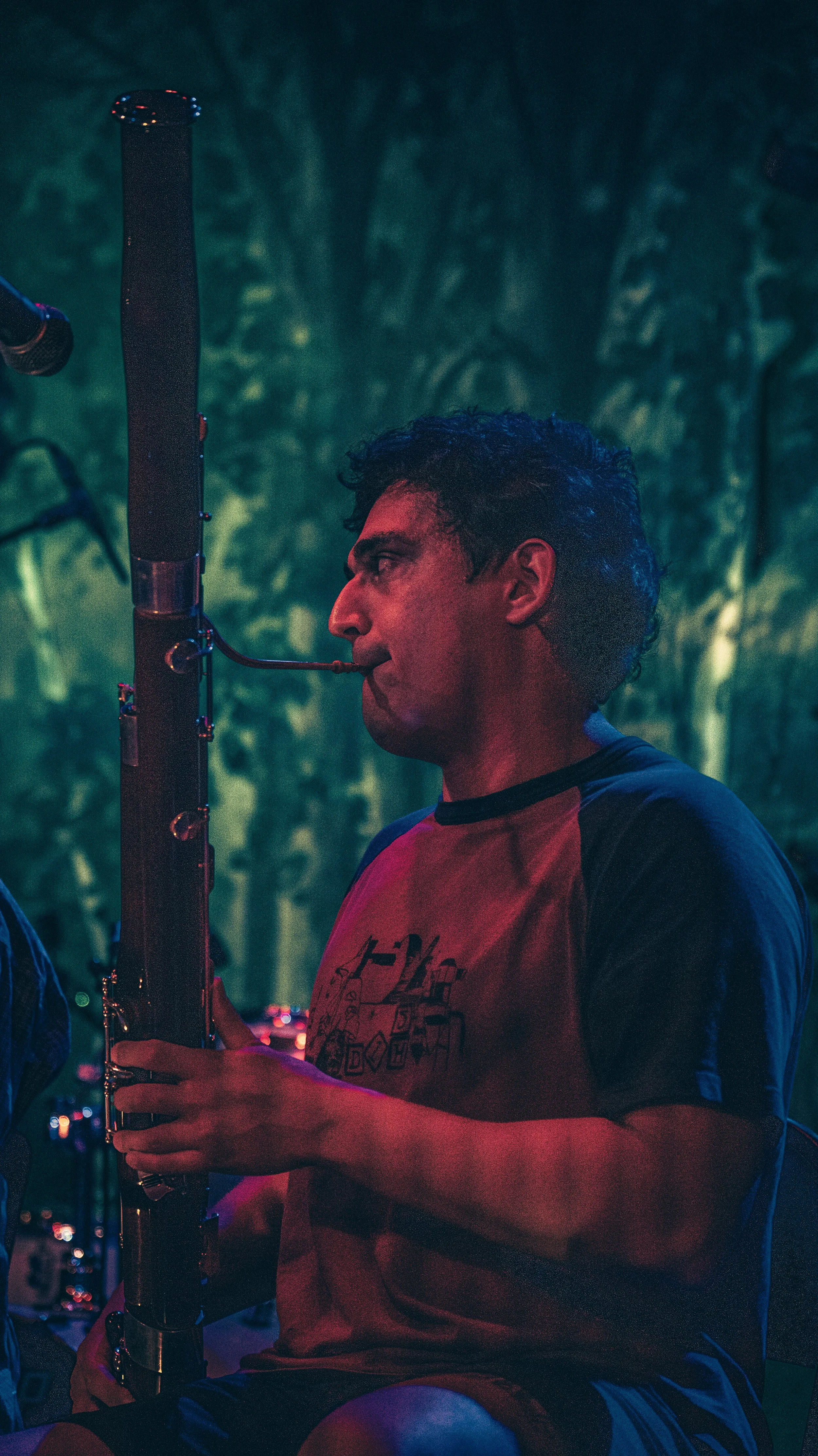 A man with short dark curly hair wearing a red and black T-shirt, playing a flute outdoors at night, with a background of trees and blurred city lights.