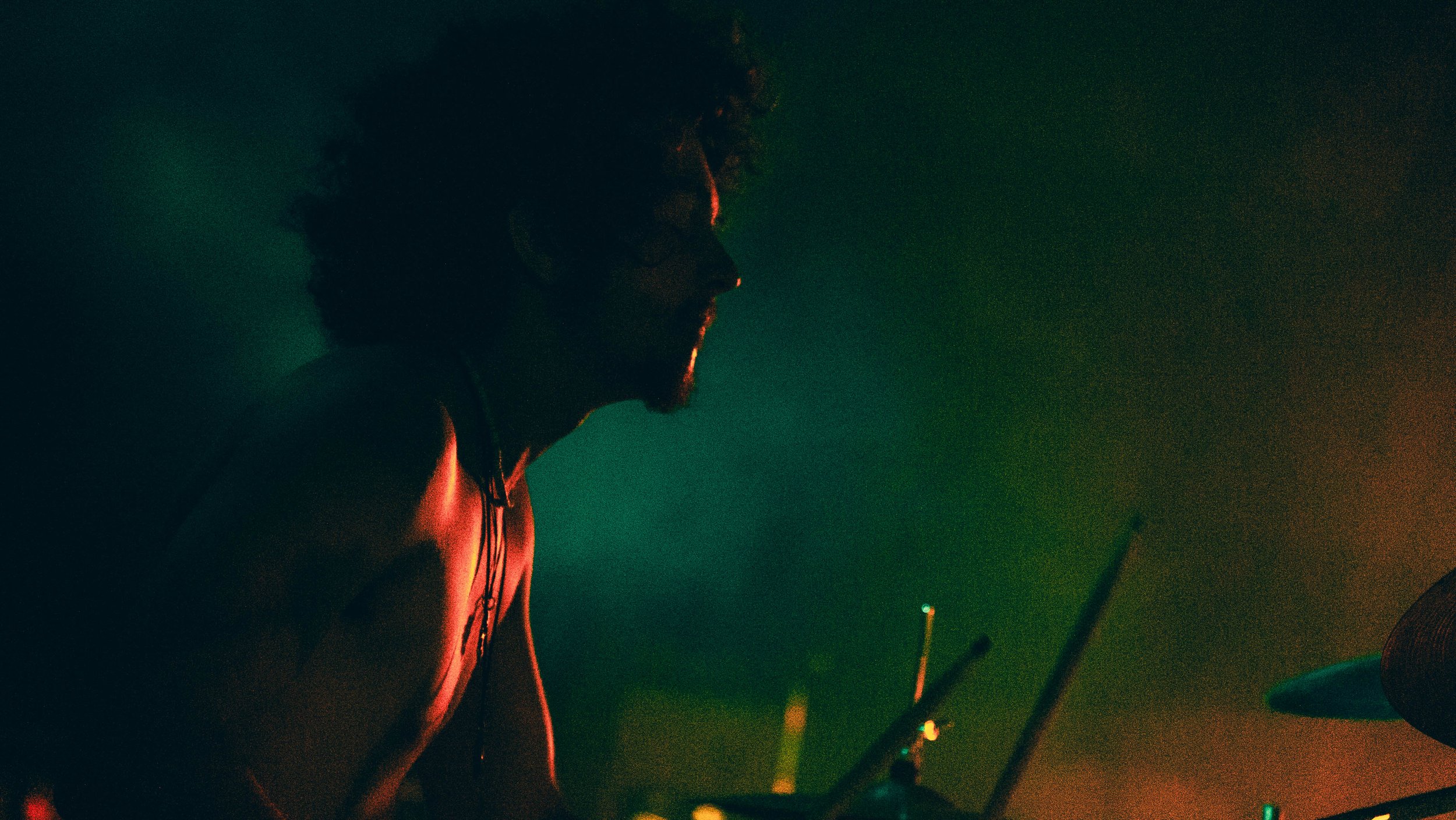 A person wearing glasses and a sleeveless top, illuminated by colorful stage lights, with a dark background and some visible equipment or structures.