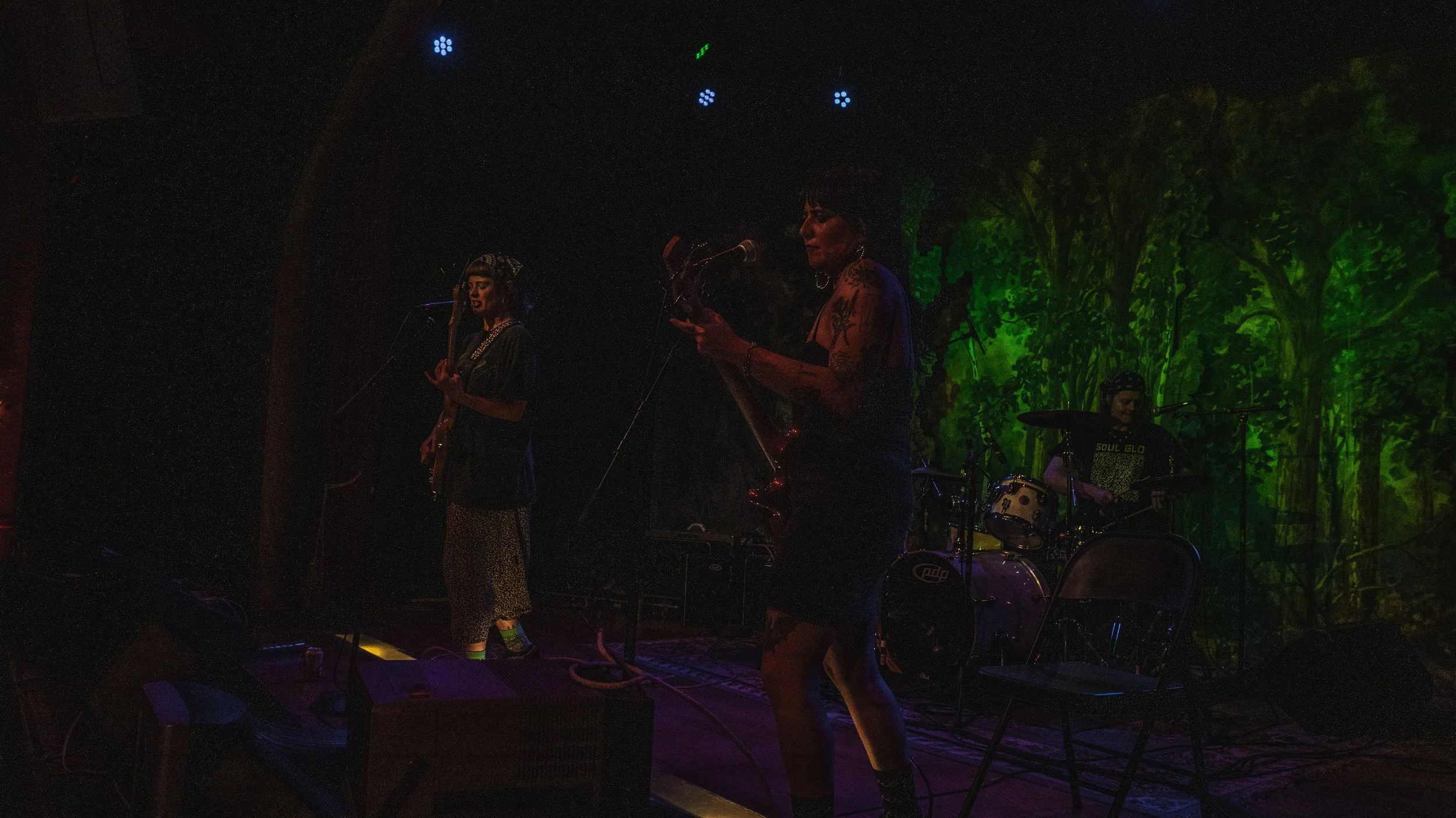A band performing on stage with three musicians, green and blue stage lighting, and a forest backdrop.