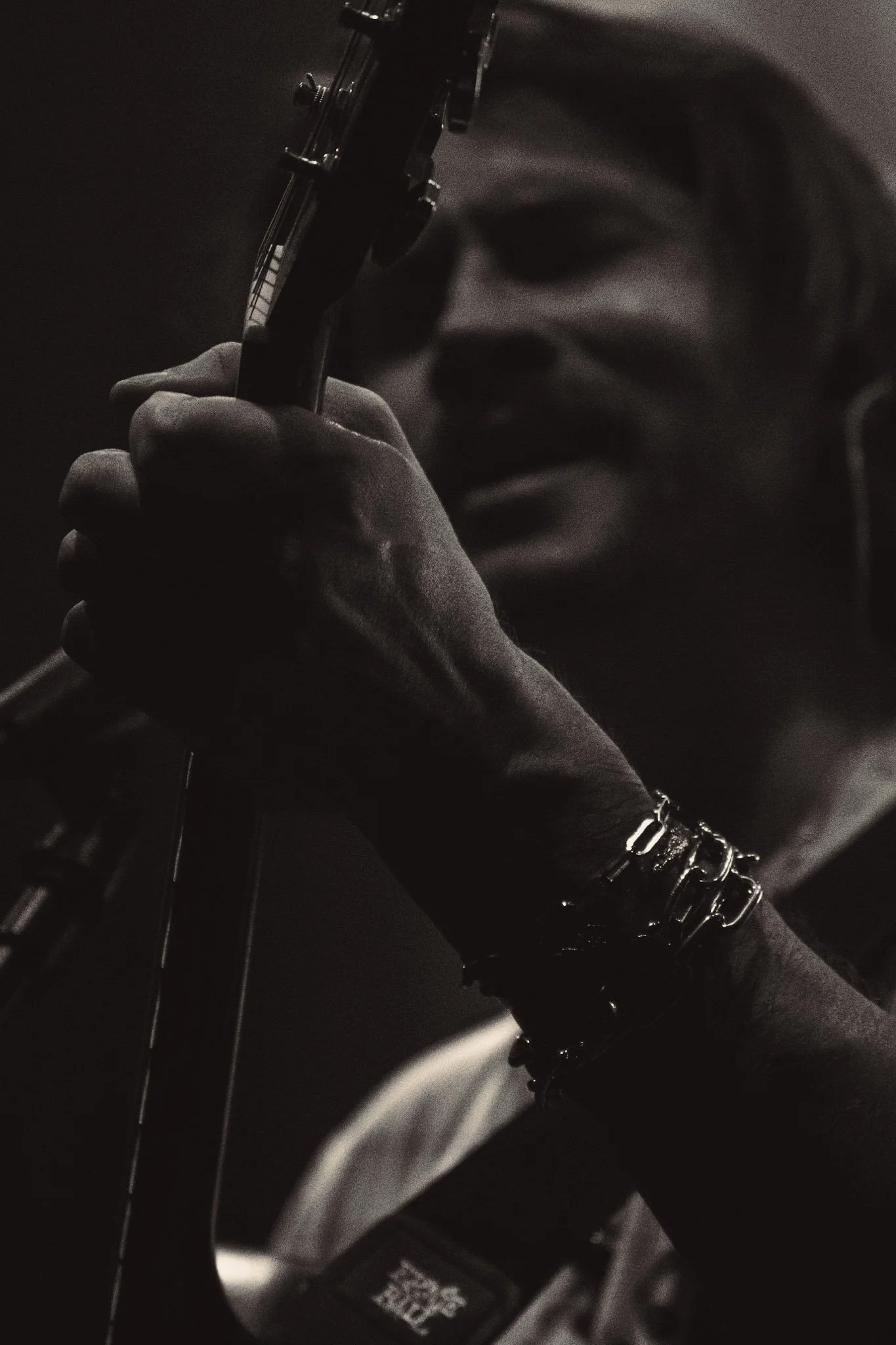 Close-up of a person's face partially visible, holding a guitar neck, wearing multiple bracelets, in a dark setting with low lighting.
