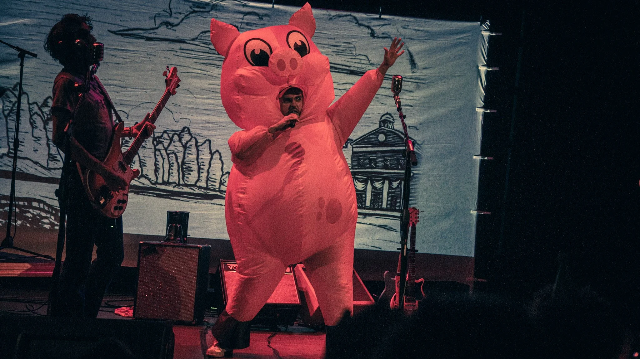 A person dressed in a pink pig costume speaking into a microphone on stage, with another person playing a guitar nearby, in front of a backdrop with sketches of buildings and trees.