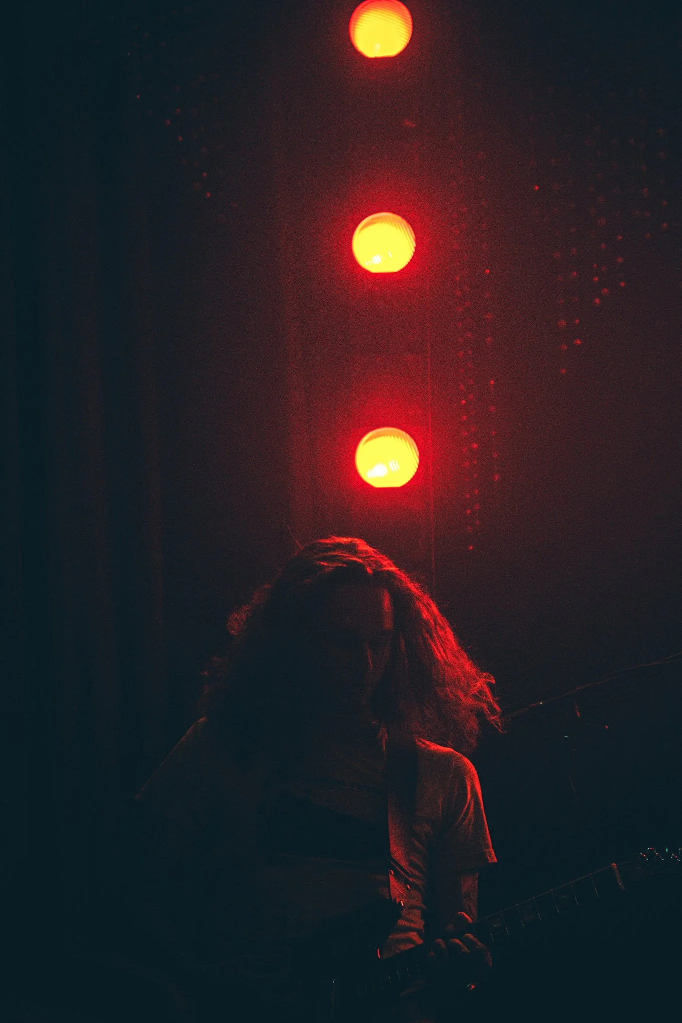 A person with long hair in front of a dark background, playing an electric guitar, illuminated by three bright red and yellow lights above.