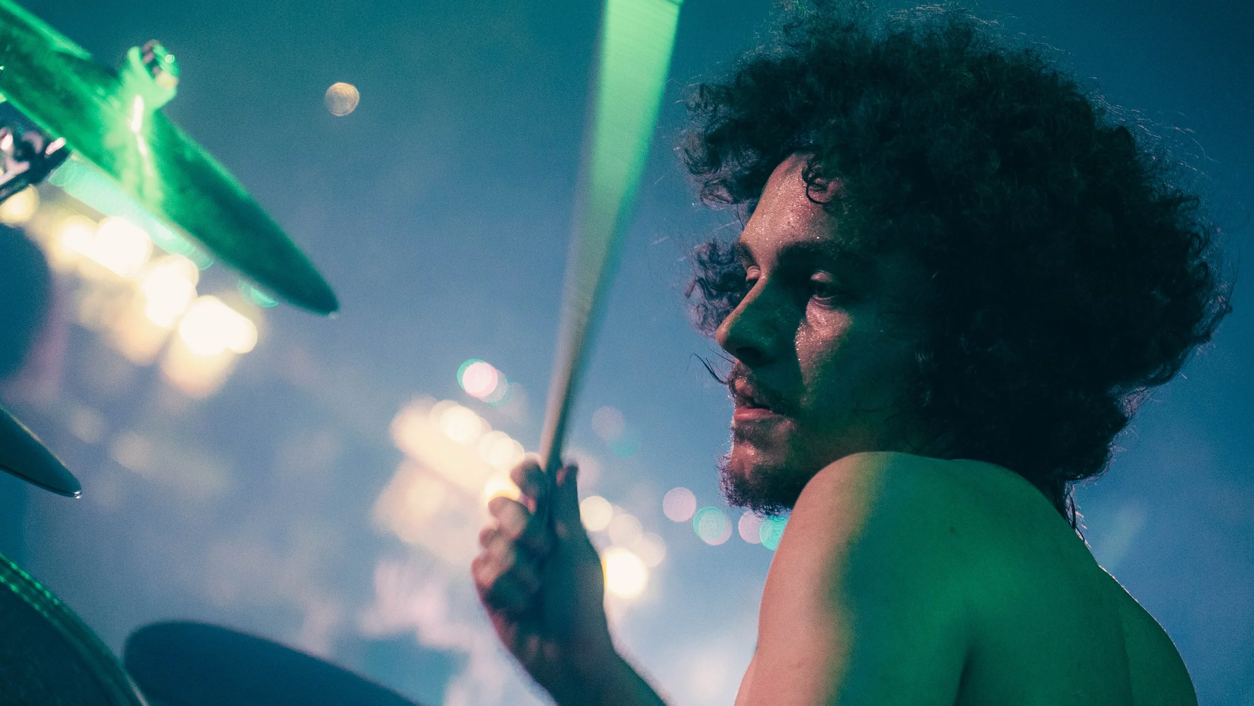 A person with curly hair playing the drums under colorful stage lights.