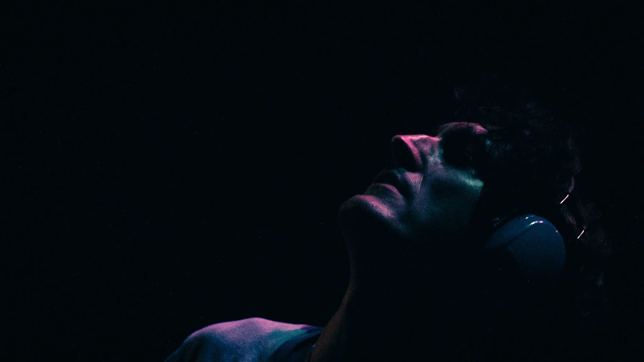 A woman with curly hair and hoop earrings, illuminated by colorful, neon-like lights, standing in a dark environment.