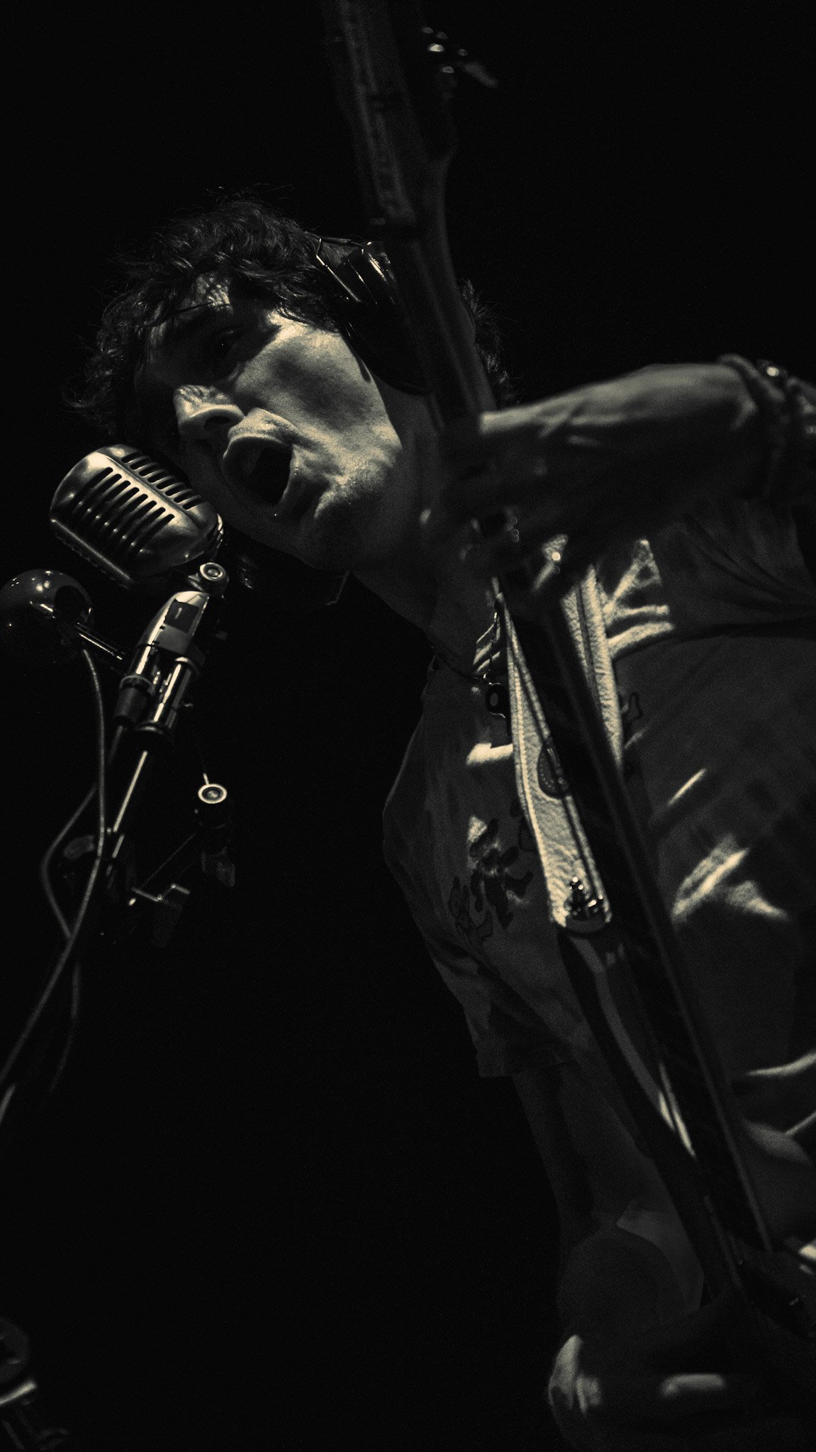 A black and white photo of a male singer with curly hair passionately singing into a vintage microphone, wearing a shiny jacket, with a dark background.