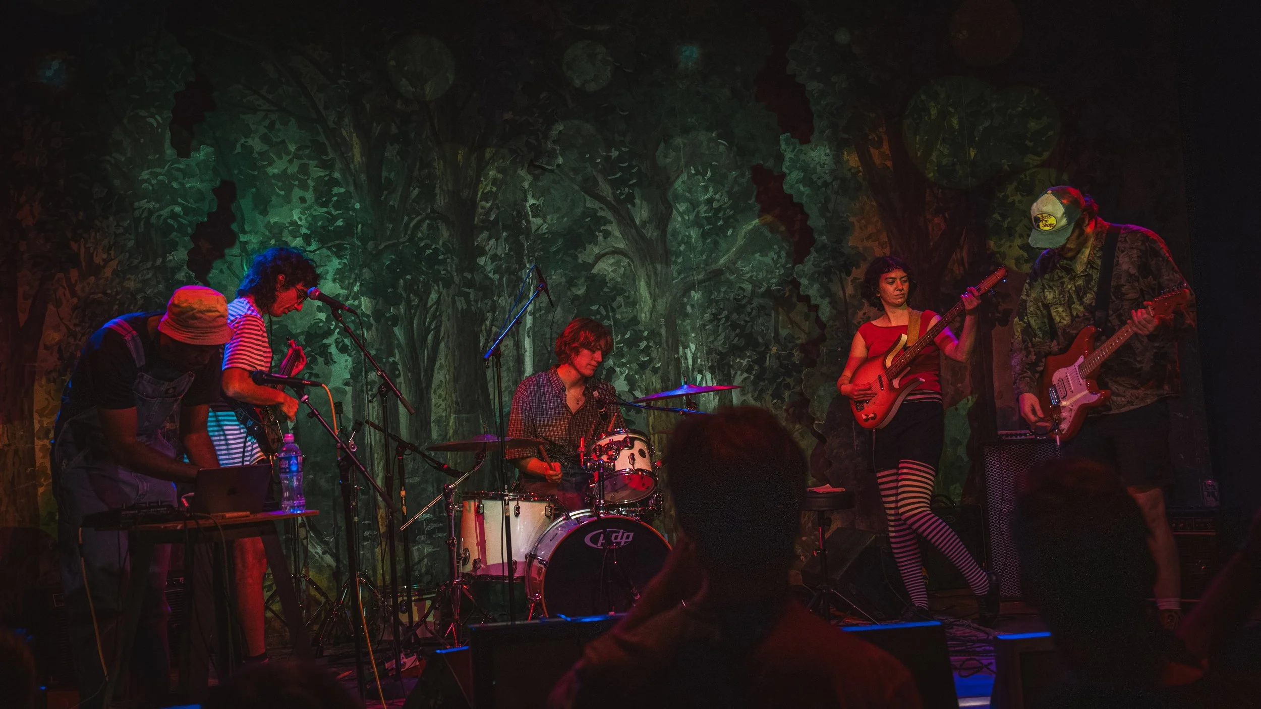 A group of musicians performing on stage with a colorful forest backdrop, including a drummer, guitarists, and vocalists, under dim stage lighting.