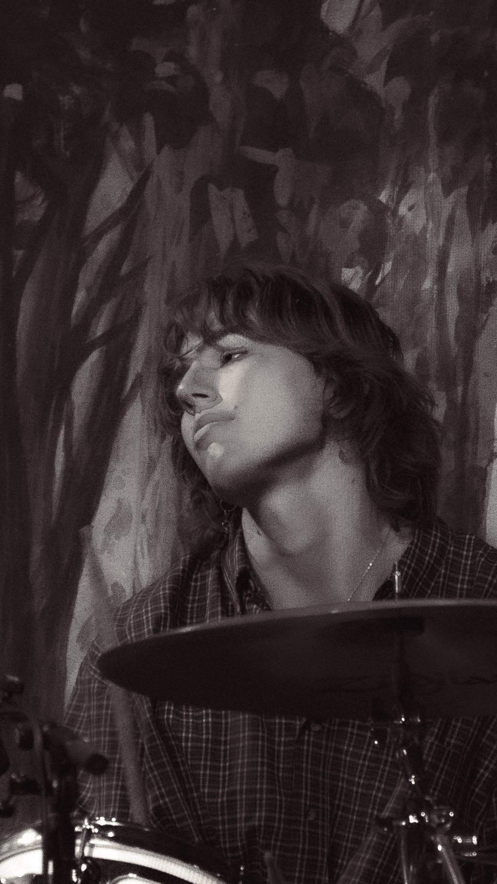A black and white photo of a young woman with short, wavy hair, wearing a checkered shirt, sitting behind a drum set, with a painted wall featuring tree branches in the background.