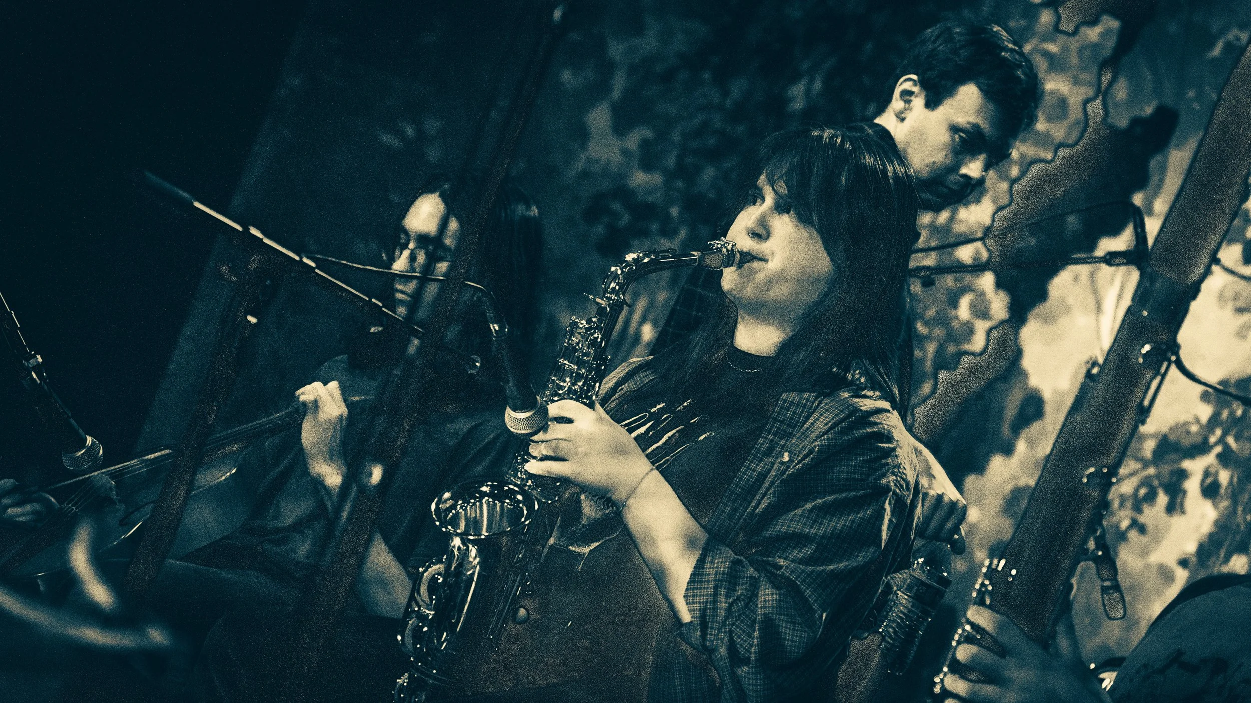 Two young adults, a woman playing a saxophone and a man holding a guitar, performing outdoors at night.