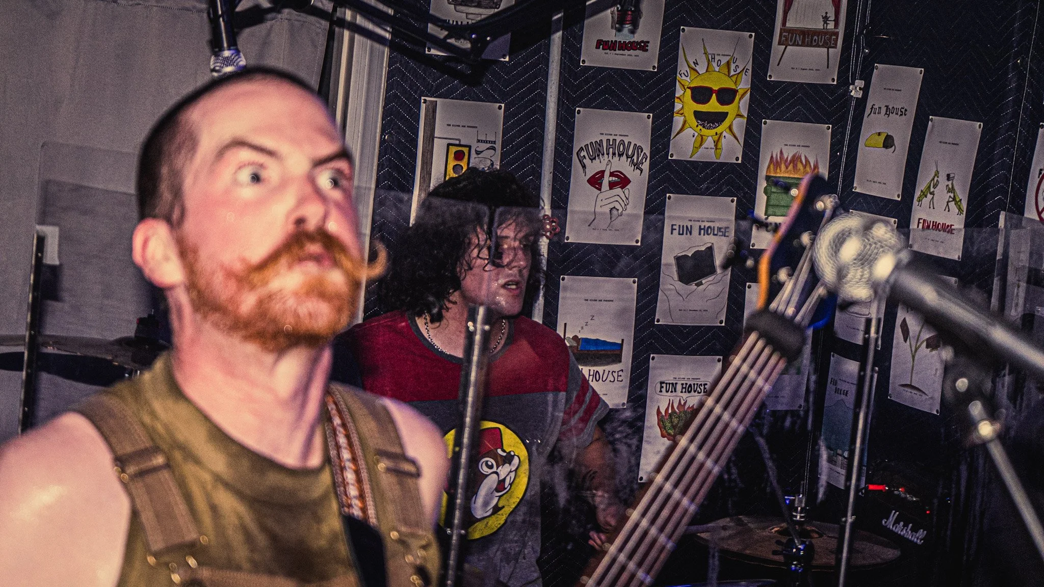 Two people are seen in a room decorated with Fun House posters, one man with a red beard in the foreground and a woman with curly dark hair in the background, near a microphone stand and musical equipment.