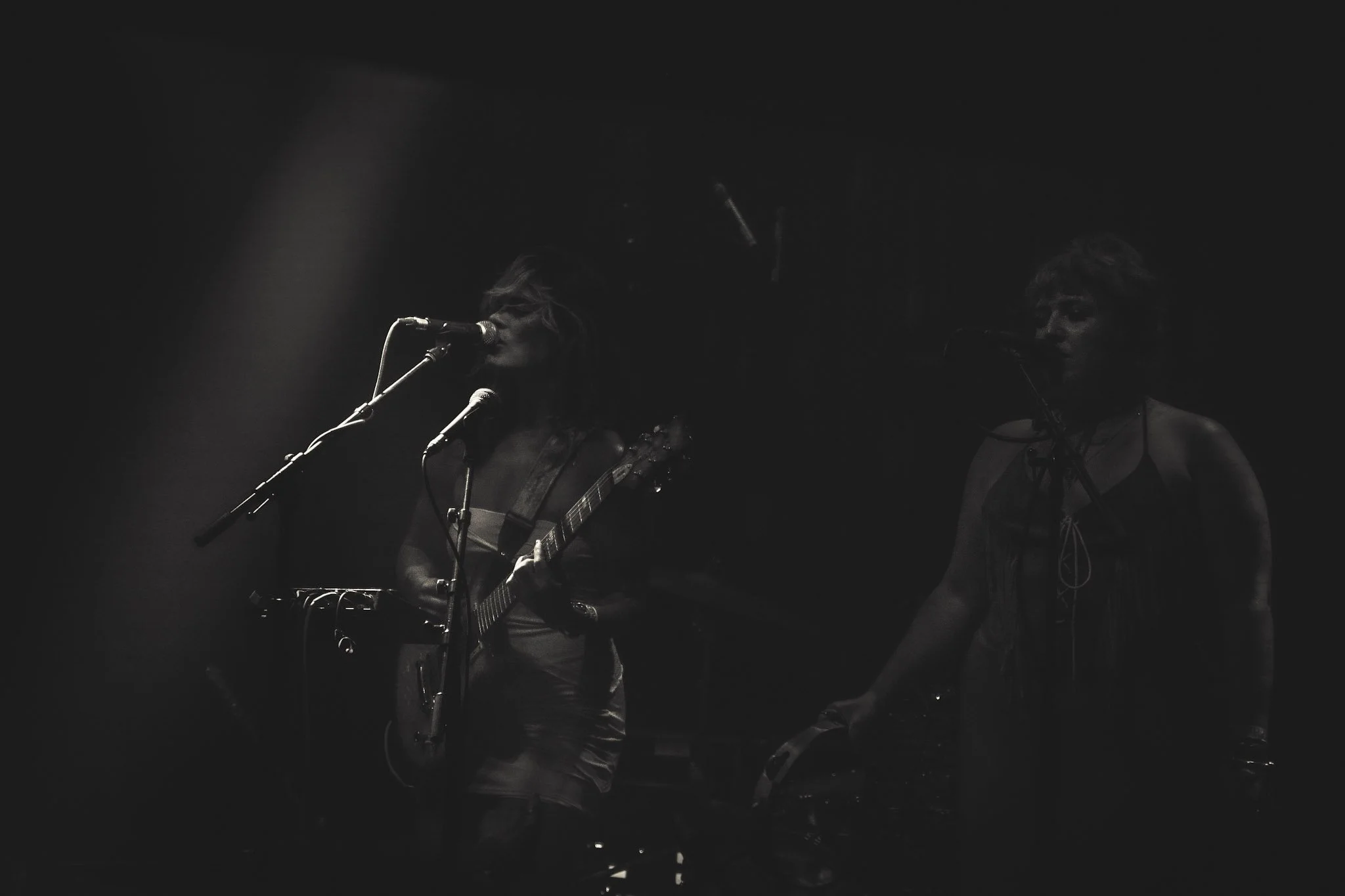 Two women performing on stage: one playing guitar, singing into a microphone, the other singing into a microphone with short hair, both dressed in dark clothing, with dark background.