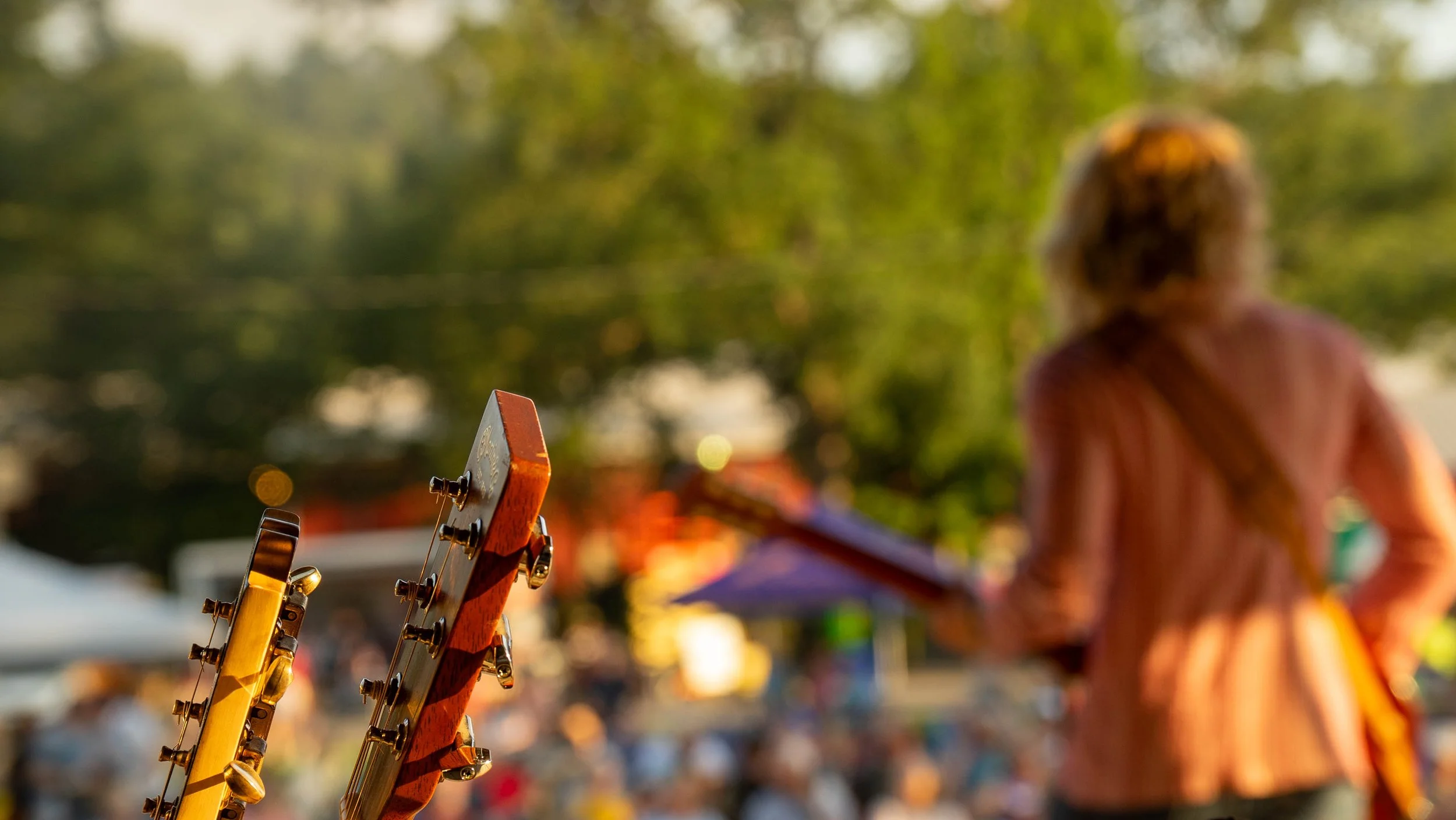 Close-up of guitar headstock with tuning pegs in focus, background blurred with a person in a plaid shirt and trees, likely at an outdoor event.