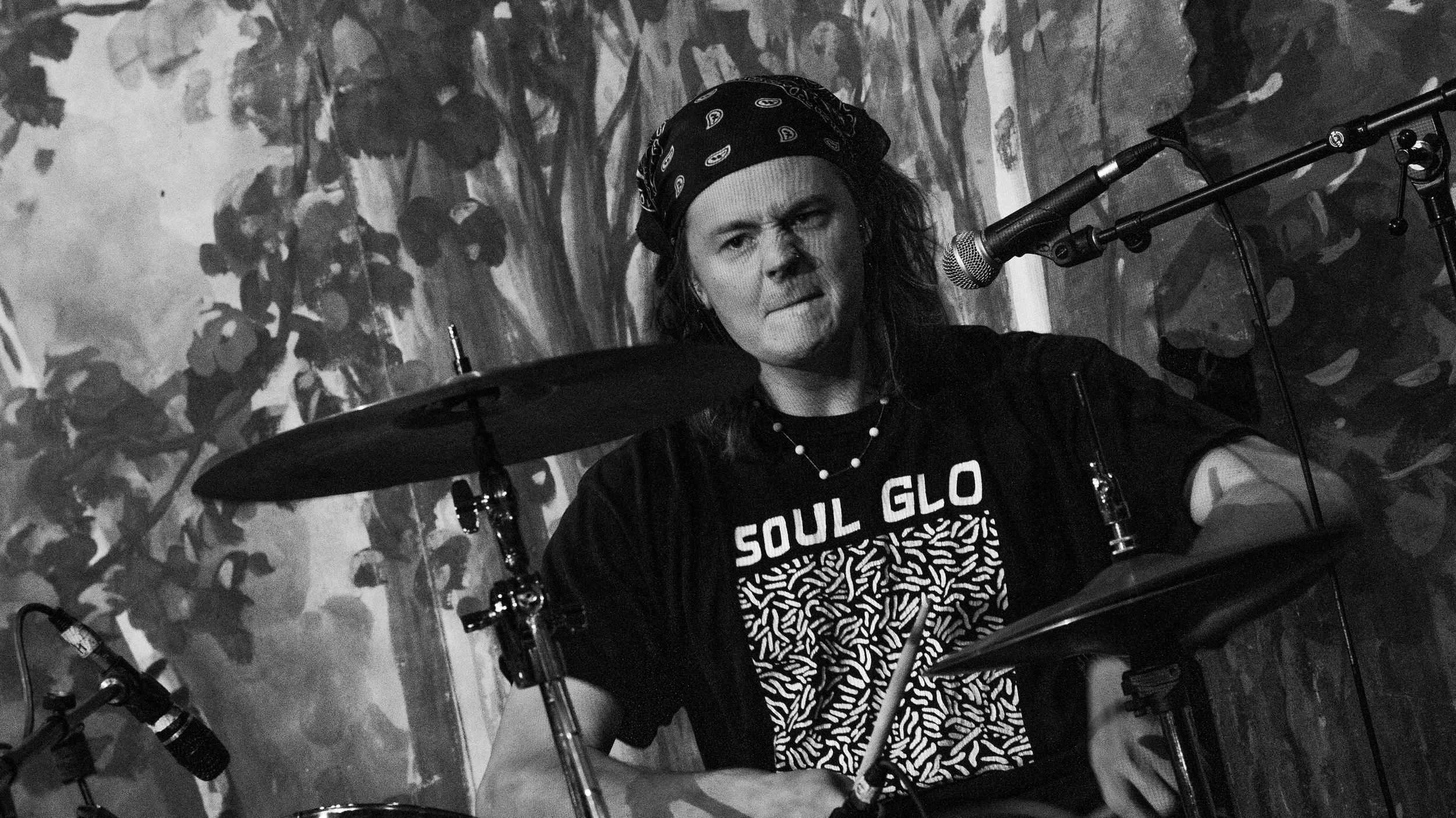 A person with long hair, wearing a bandana on their head and a t-shirt that reads 'SOUL GLO,' appears to be performing as a drummer with a drum kit. The person is biting their lip and looking intensely forward, with a microphone set up in front of th