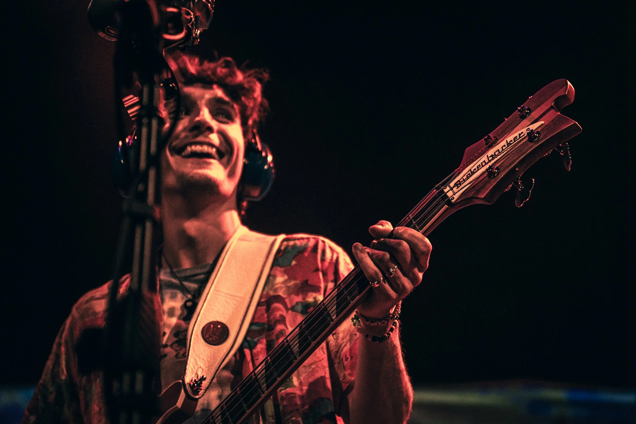 A person with curly hair smiling and playing an acoustic guitar at night, wearing a colorful patterned shirt and bracelets, with a dark background.