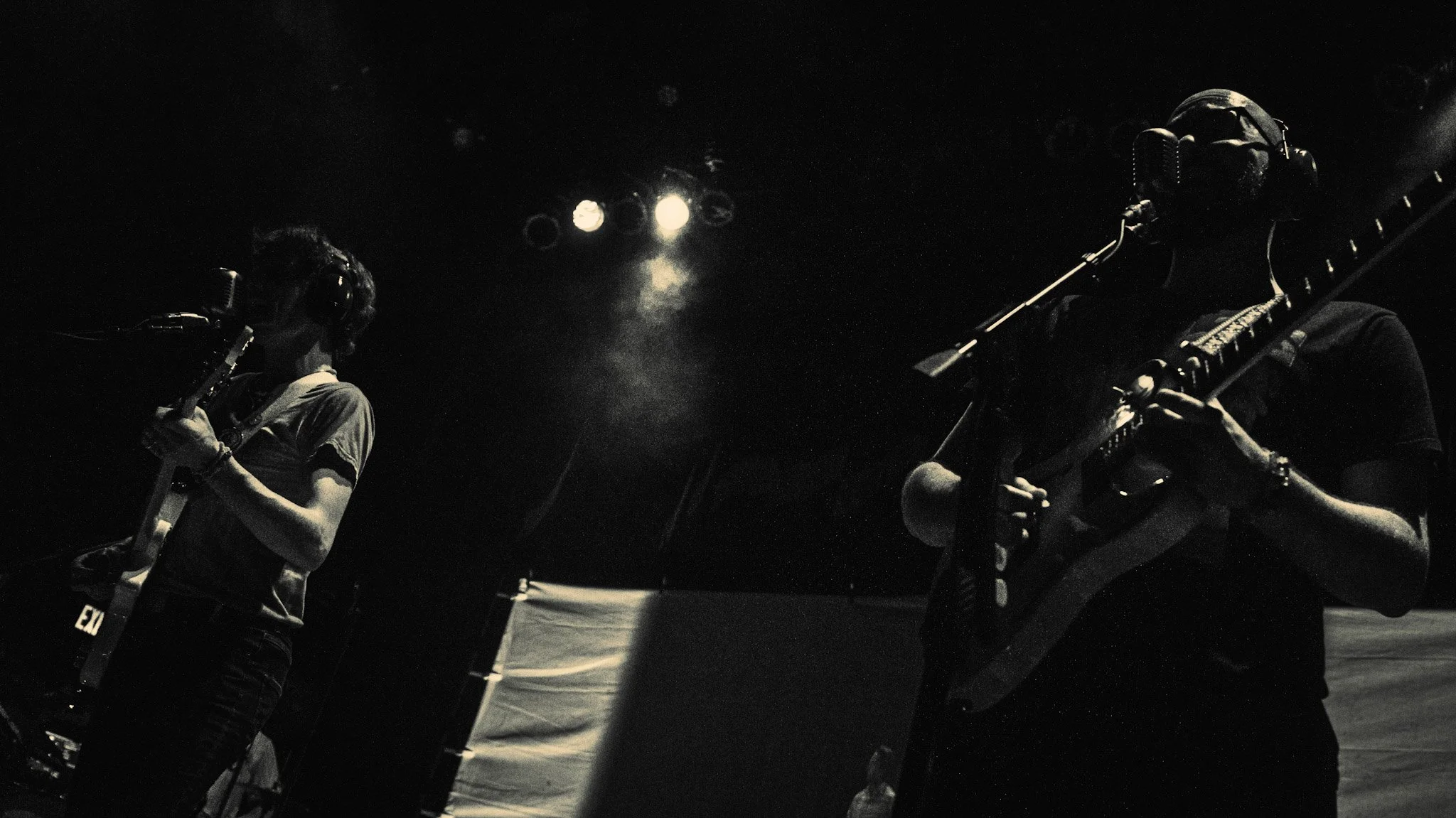 Two musicians wearing headphones and playing guitars on stage at night, with bright stage lights in the background.