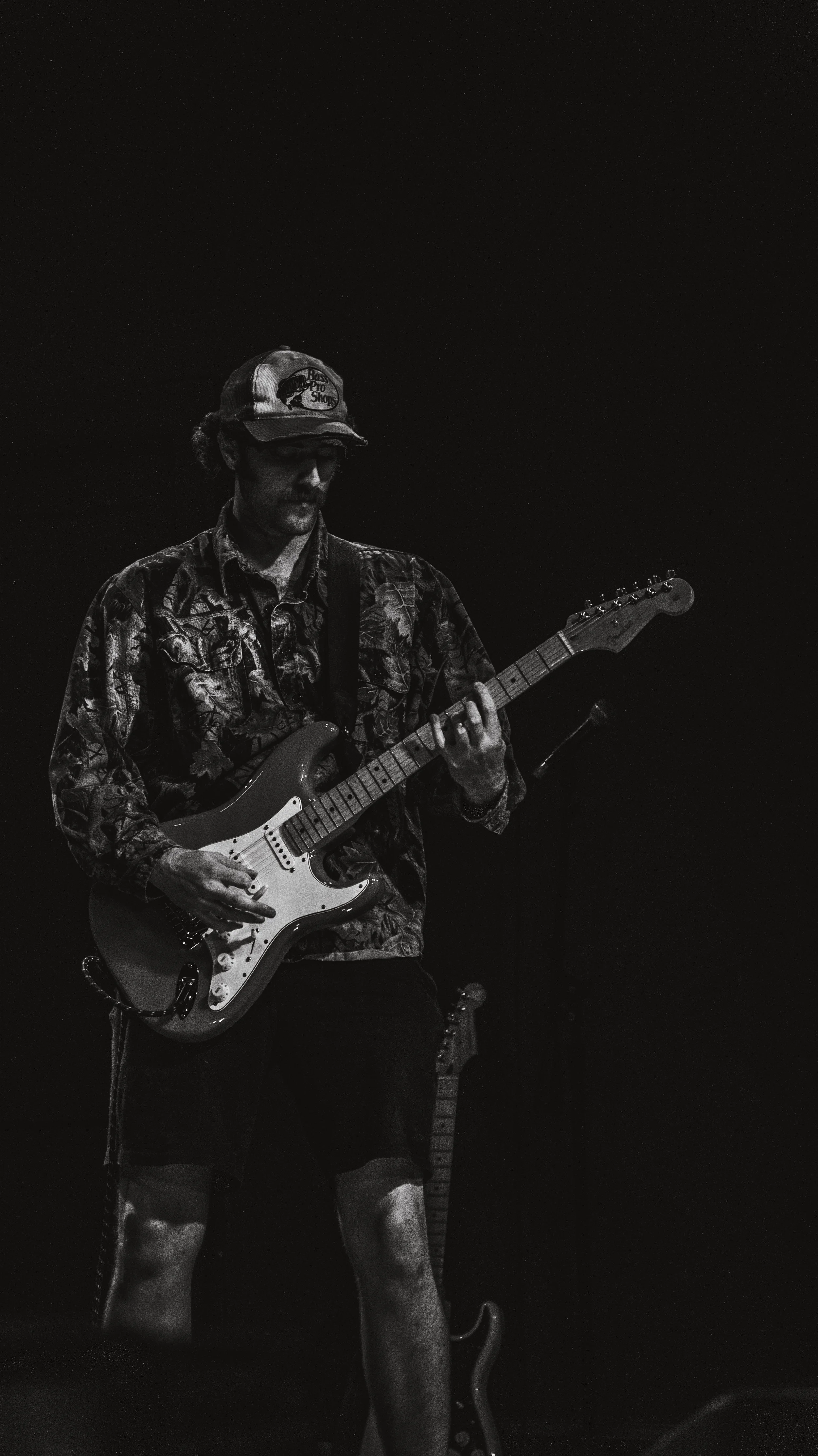 A man wearing a camouflage jacket, shorts, and a cap with a bass pro shop logo, playing an electric guitar on stage in a dark setting.