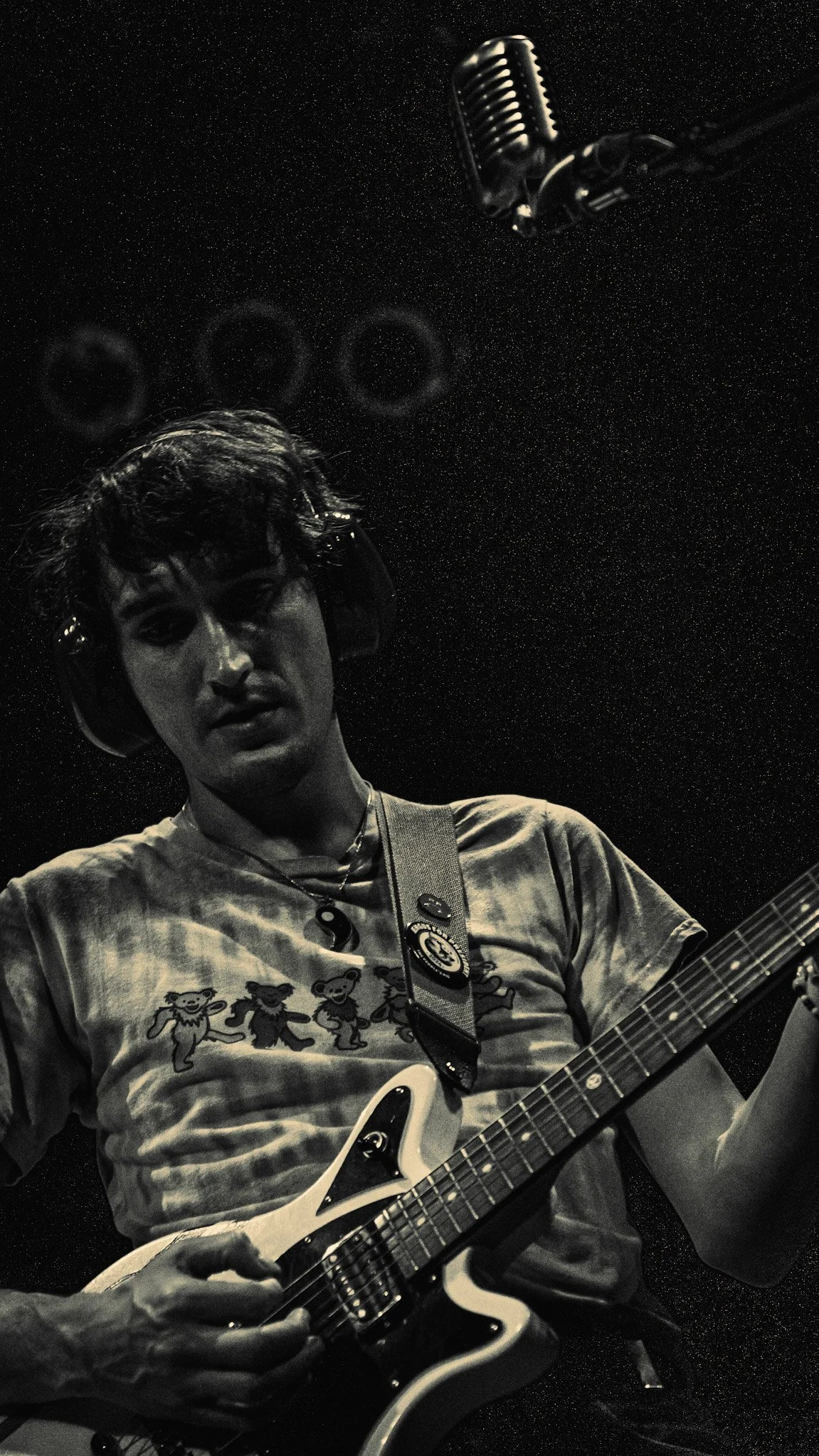 A young man with curly hair wearing glasses, headphones, and a T-shirt with teddy bear graphics, playing an electric guitar in a dimly lit room with a starry sky background and a vintage microphone overhead.
