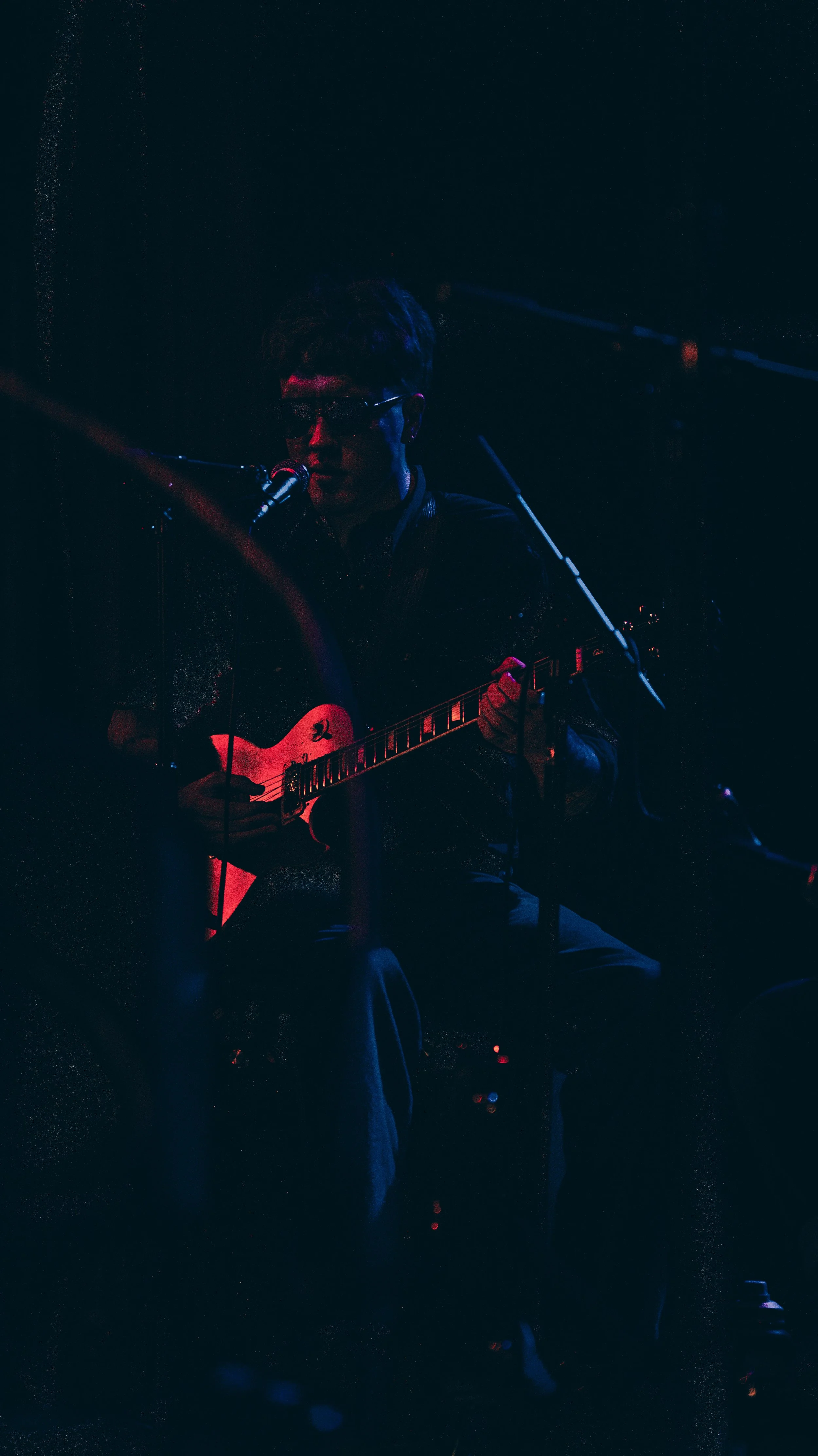 A musician playing a guitar and singing into a microphone on a dark stage, illuminated by dim stage lights.