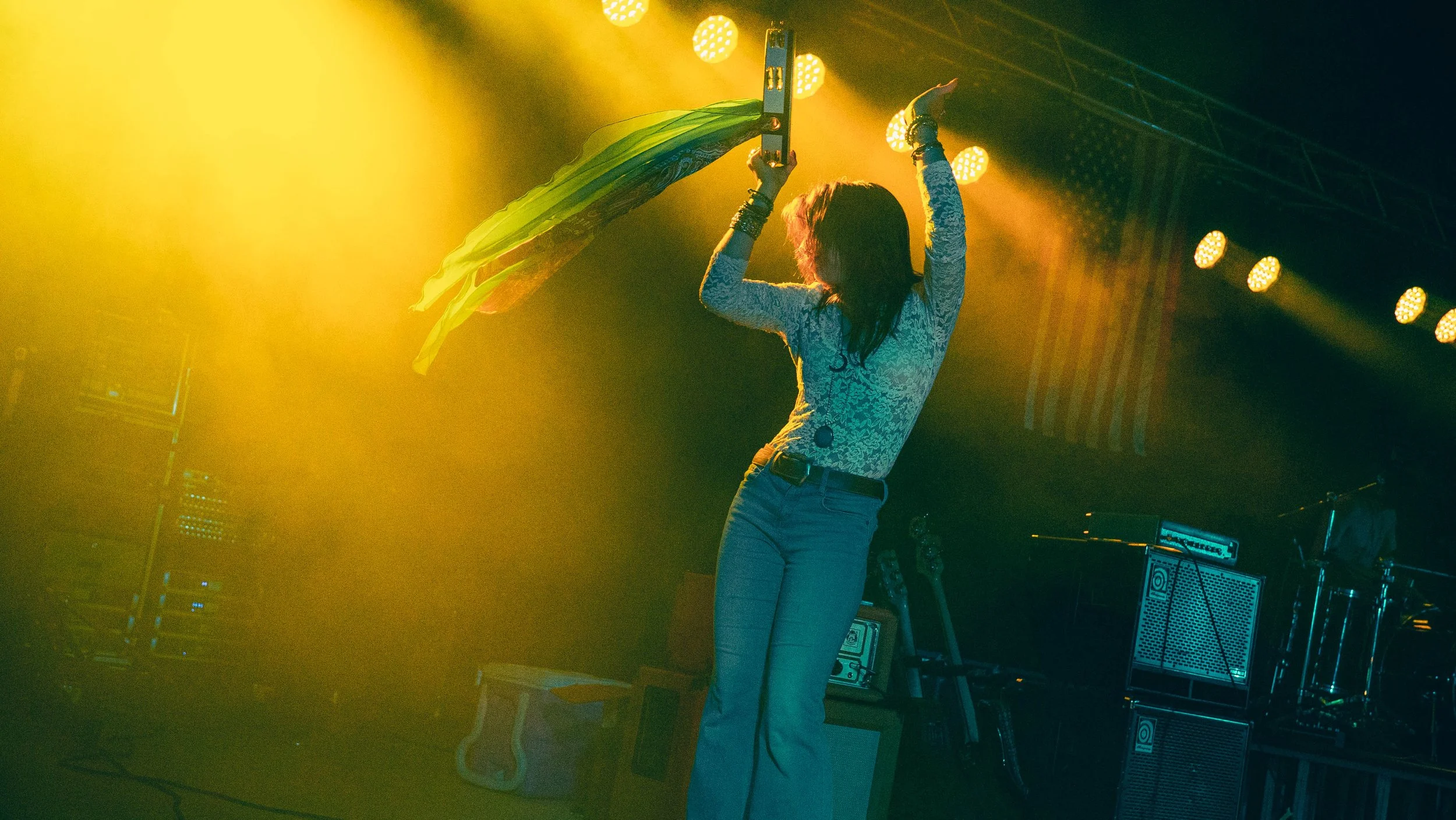 A woman on stage singing into a microphone, holding a green and yellow fabric, with stage lights shining and audio equipment in the background.