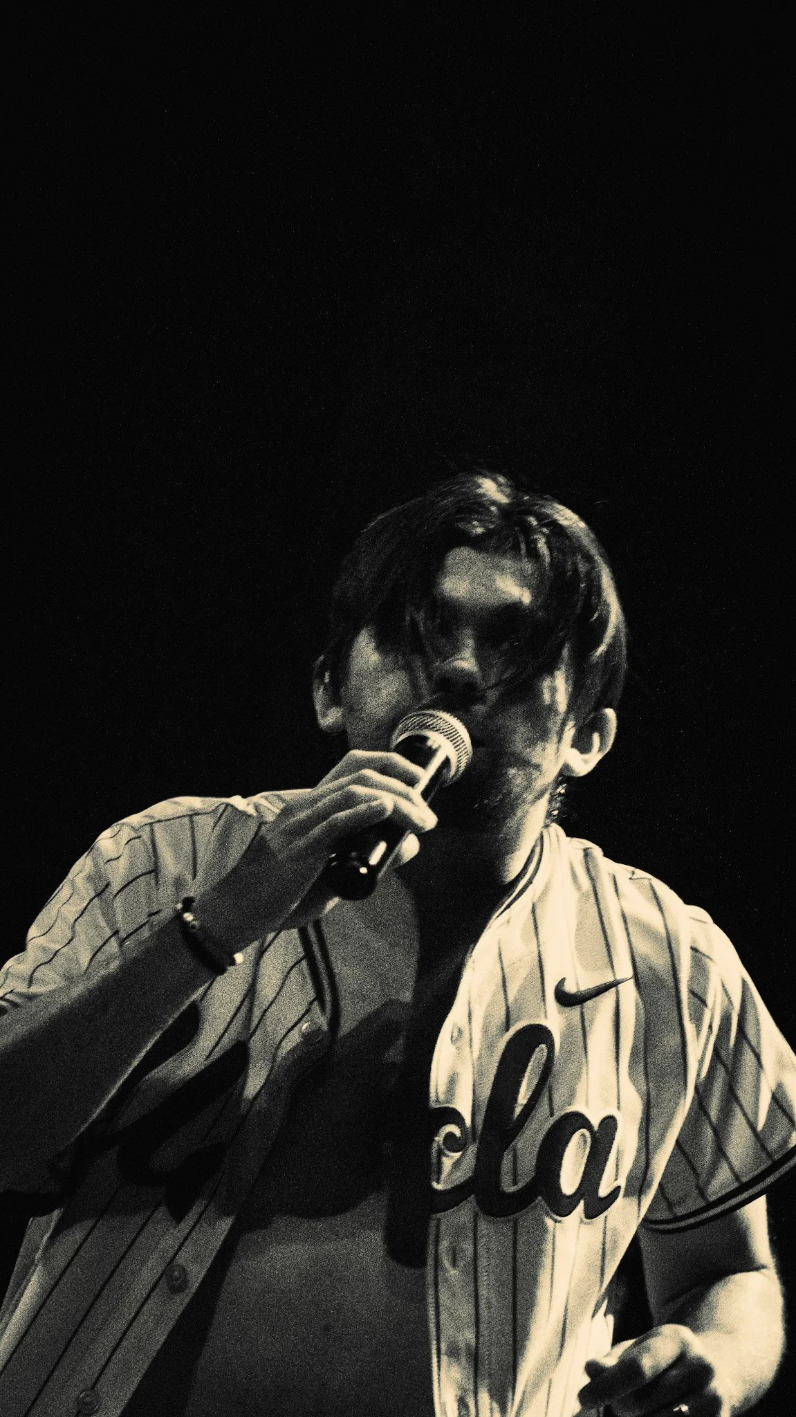 A man wearing a baseball jersey and holding a microphone, appears to be singing or speaking, with dark background.