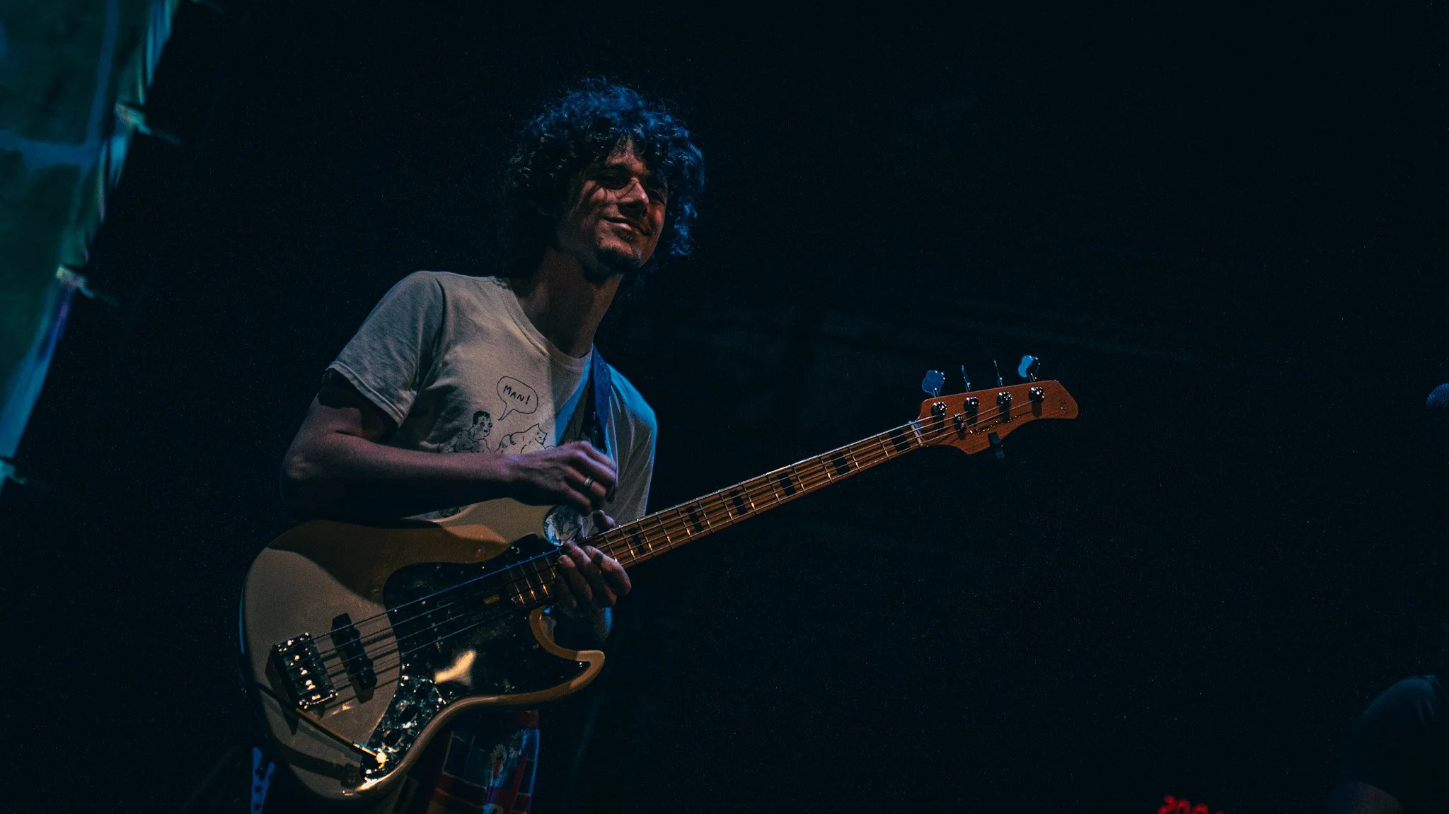 A person with curly dark hair playing an electric bass guitar on stage during a performance, illuminated by dim blue and red lighting.