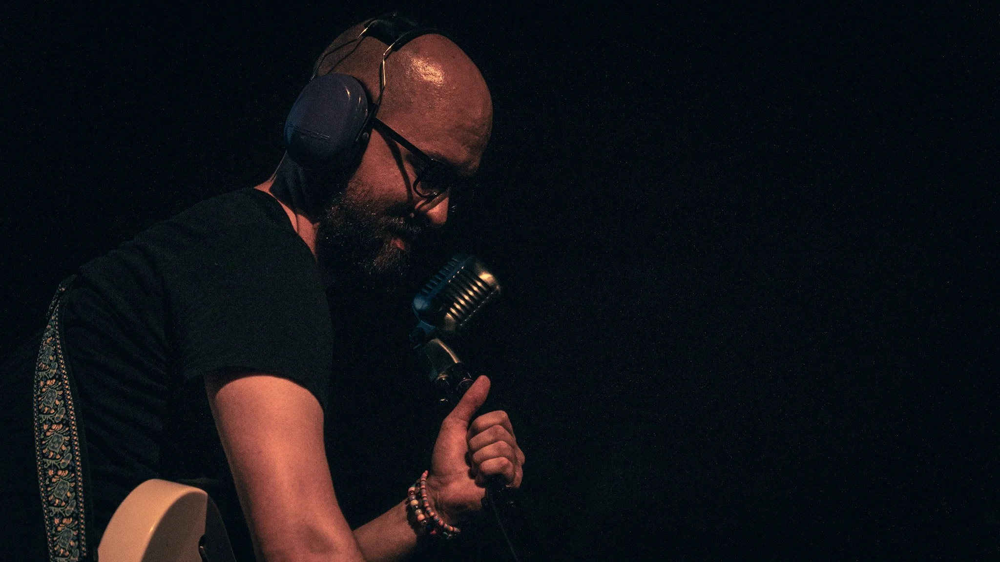 A man wearing headphones, glasses, and a black shirt, singing into a retro microphone, holding a guitar, in a dark setting.