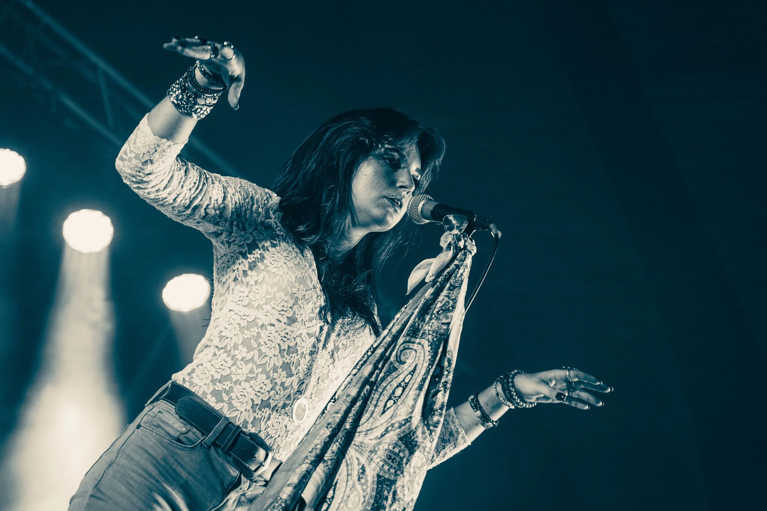 A female performer singing on stage into a microphone, dressed in a lace top with accessories on her wrist, with stage lights in the background.