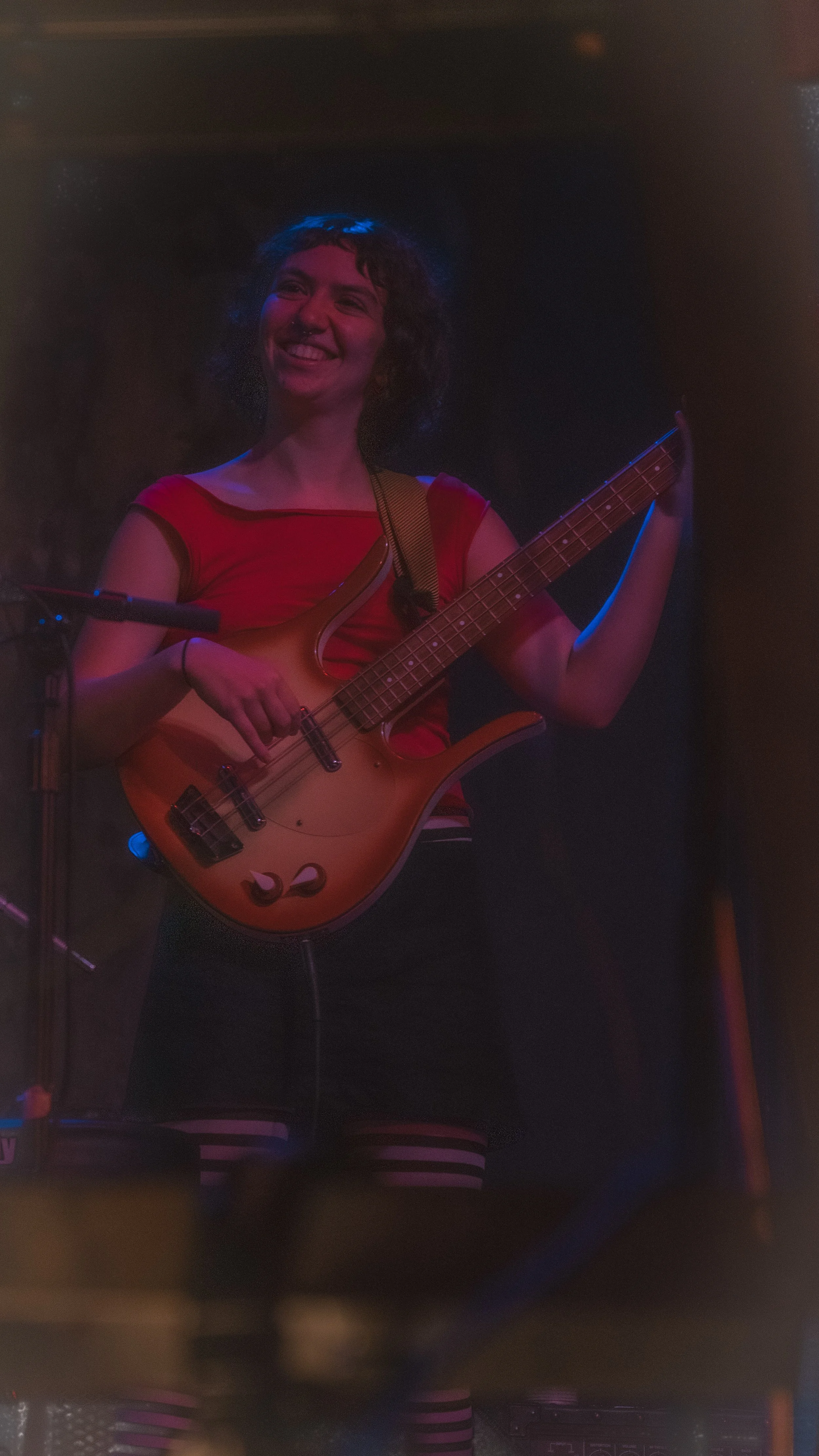 A woman with curly hair smiling and playing a bass guitar on stage, wearing a red top and striped shorts, with stage lighting creating a dark background.