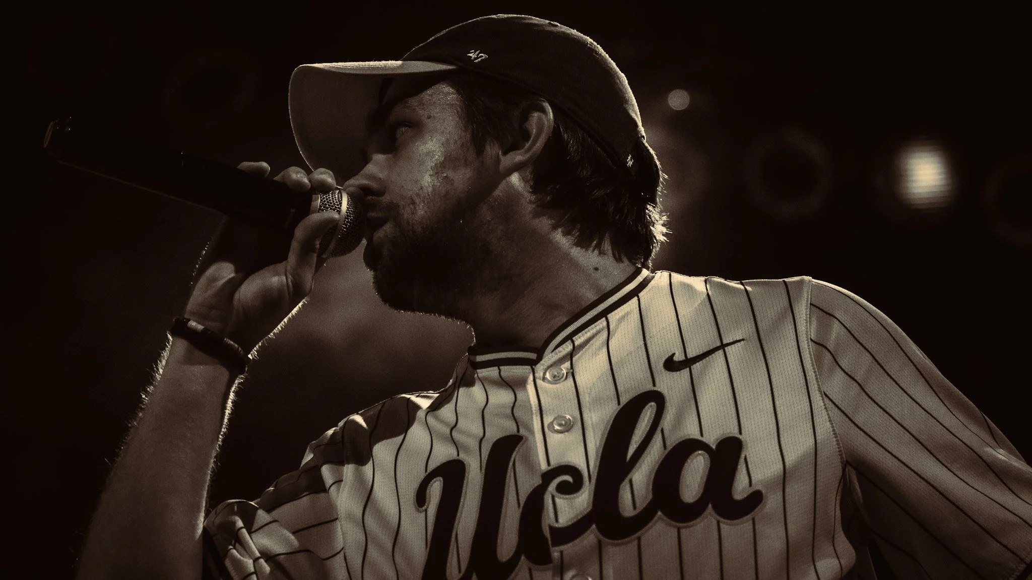 A man in a baseball uniform and cap is singing into a microphone during a performance.