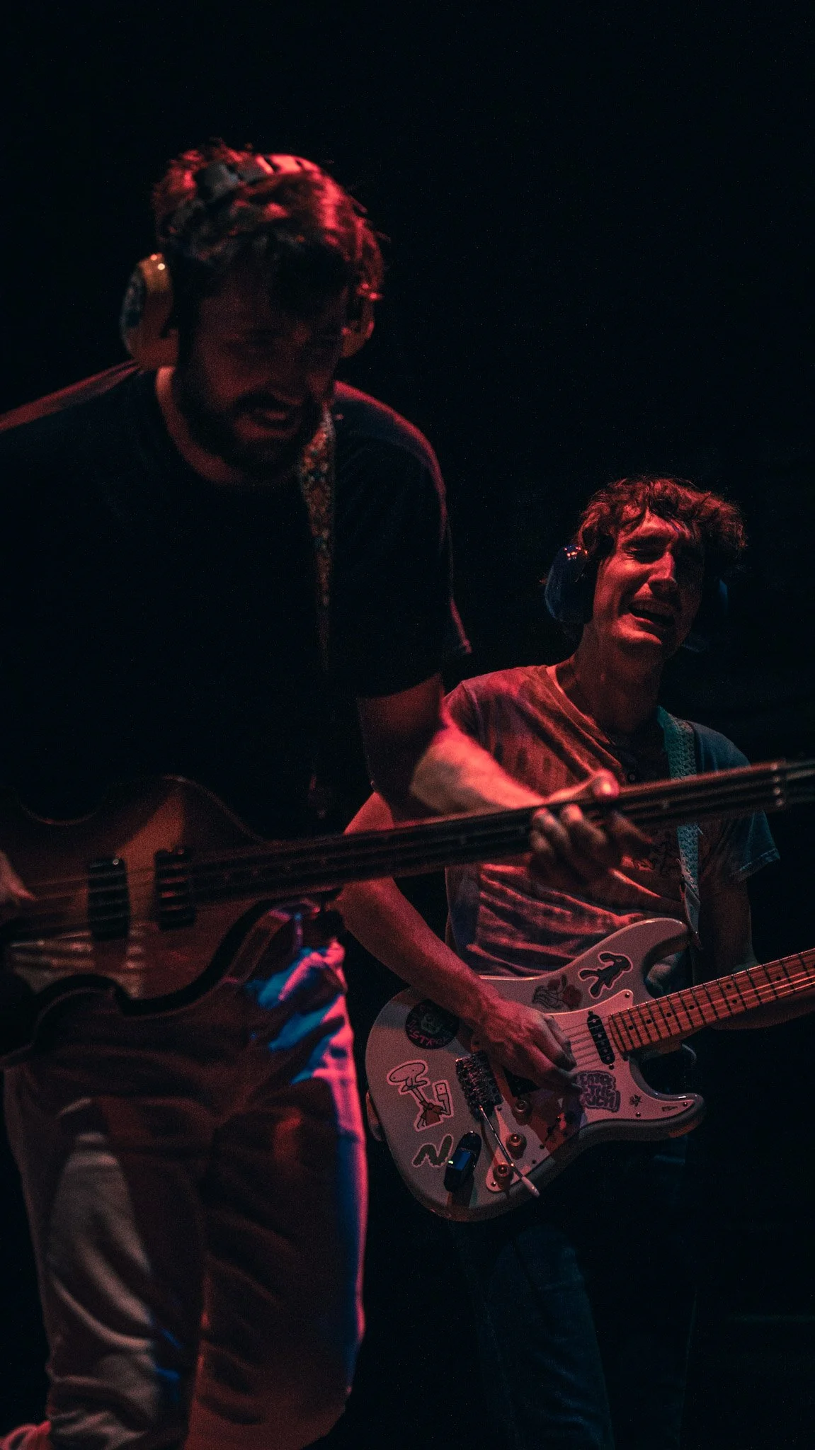 Two musicians performing on stage at night, one playing an acoustic guitar and the other playing an electric guitar, both wearing headphones.