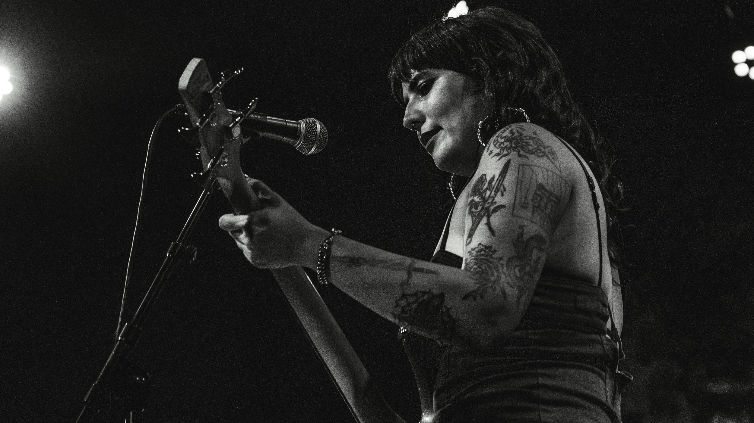 Black and white photo of a female musician playing guitar and singing into a microphone on stage, with stage lights in the background.