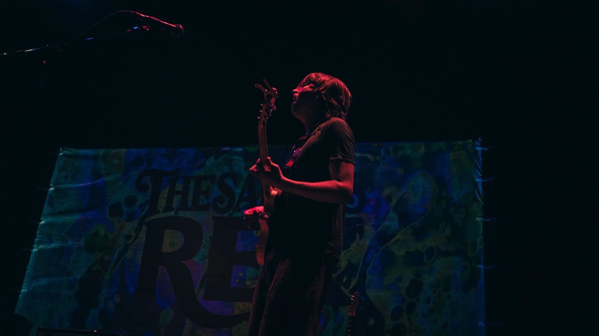 A person playing guitar on stage with a colorful banner in the background, illuminated by dark stage lighting.