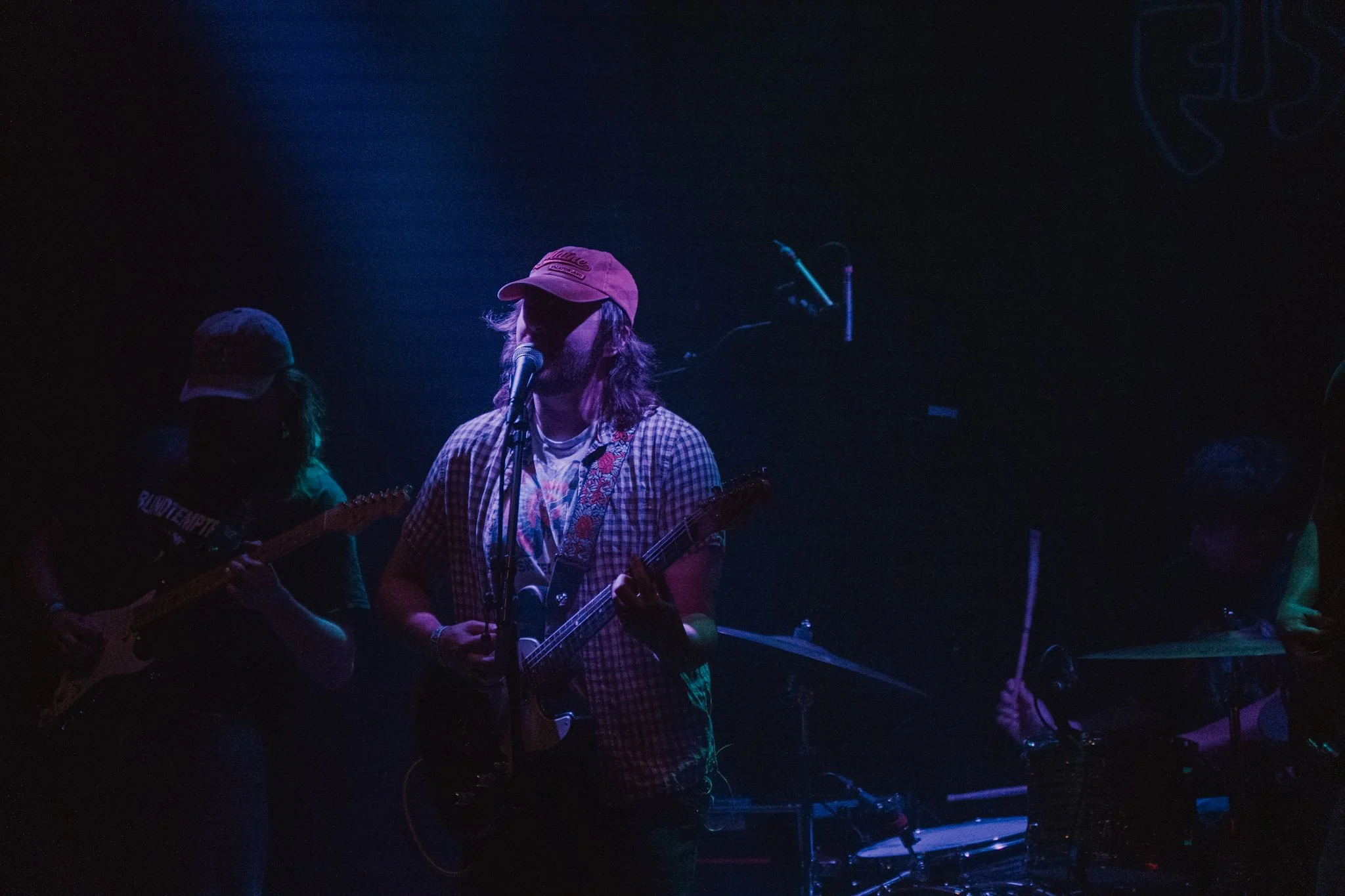 Musicians performing on stage in a dimly lit setting, one with a pink cap singing into a microphone and playing guitar, with others around playing instruments.