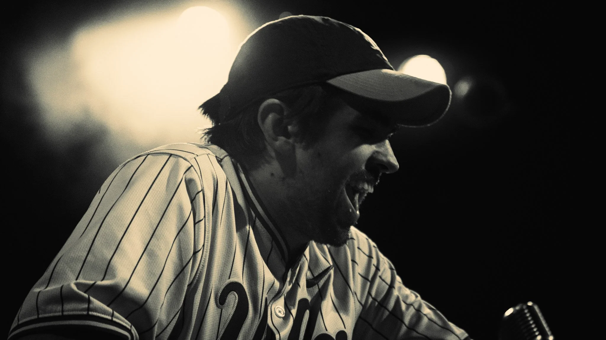 A person wearing a baseball cap and sunglasses, smiling while wearing a pinstripe sports jersey.