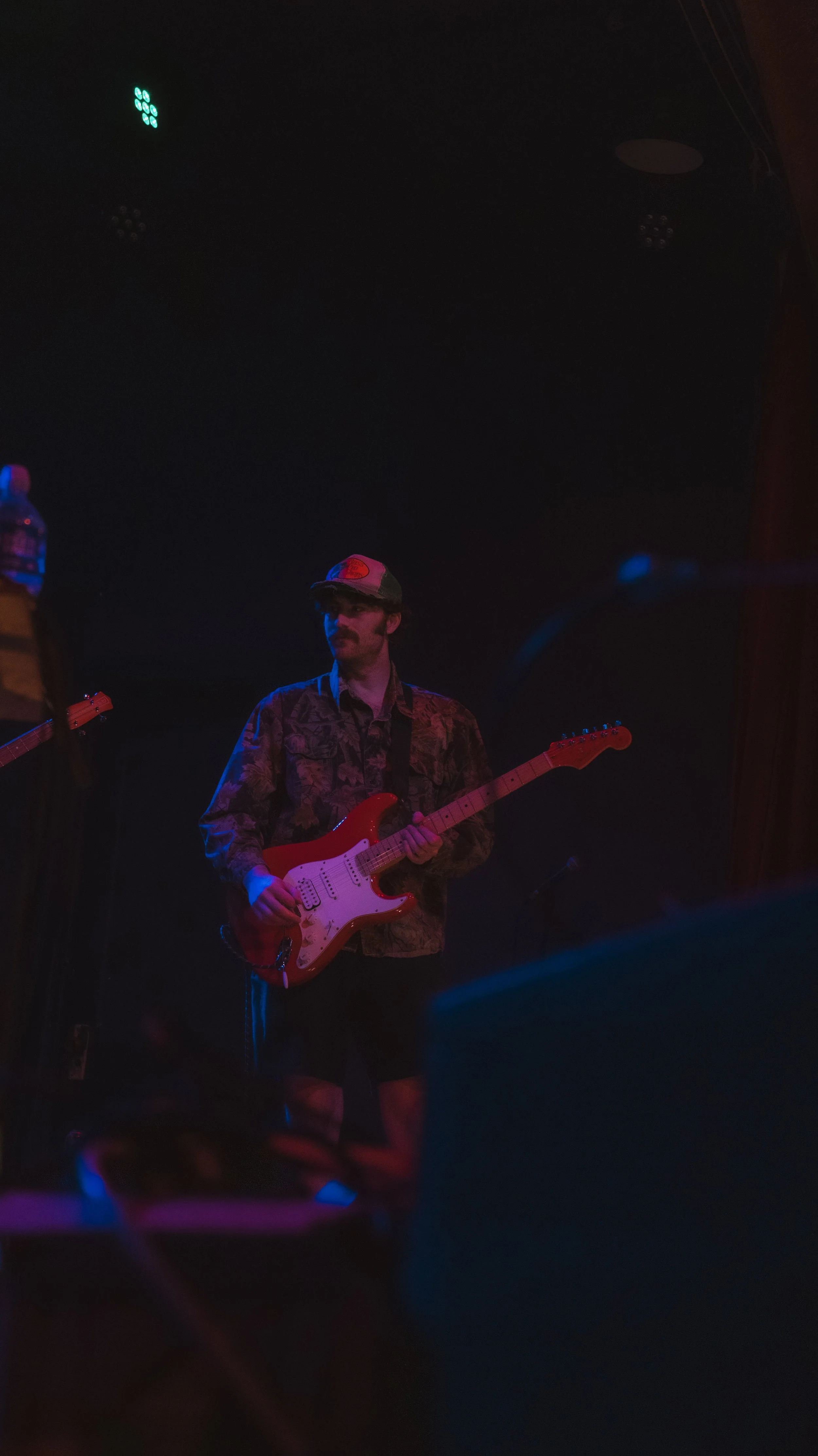 A musician playing an electric guitar on stage under dim lighting with blue and red hues, wearing a cap and a patterned shirt.