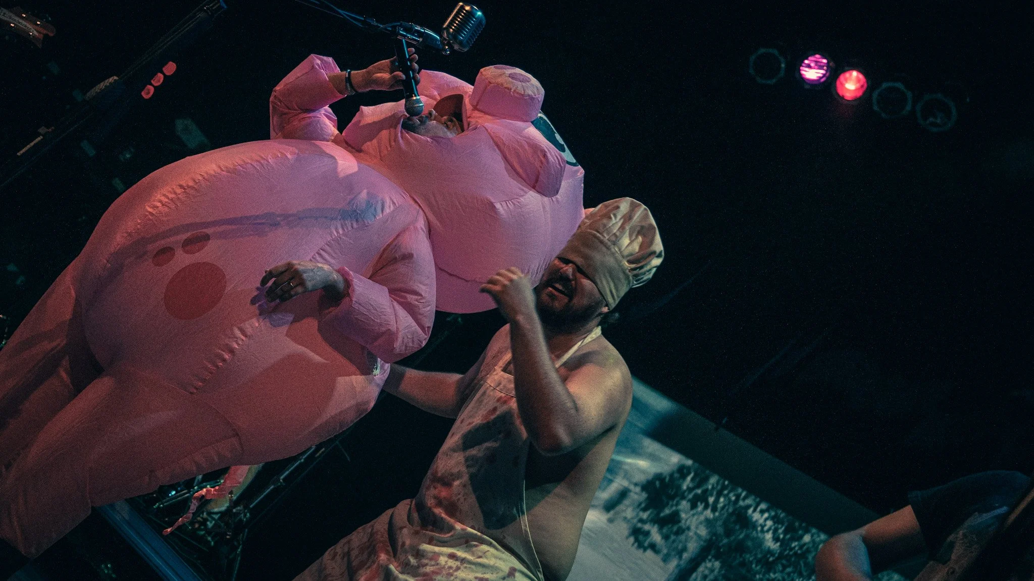Person in pink inflatable costume performing onstage with a microphone, wearing a head covering, and another person in beige clothing gesturing with their hand.