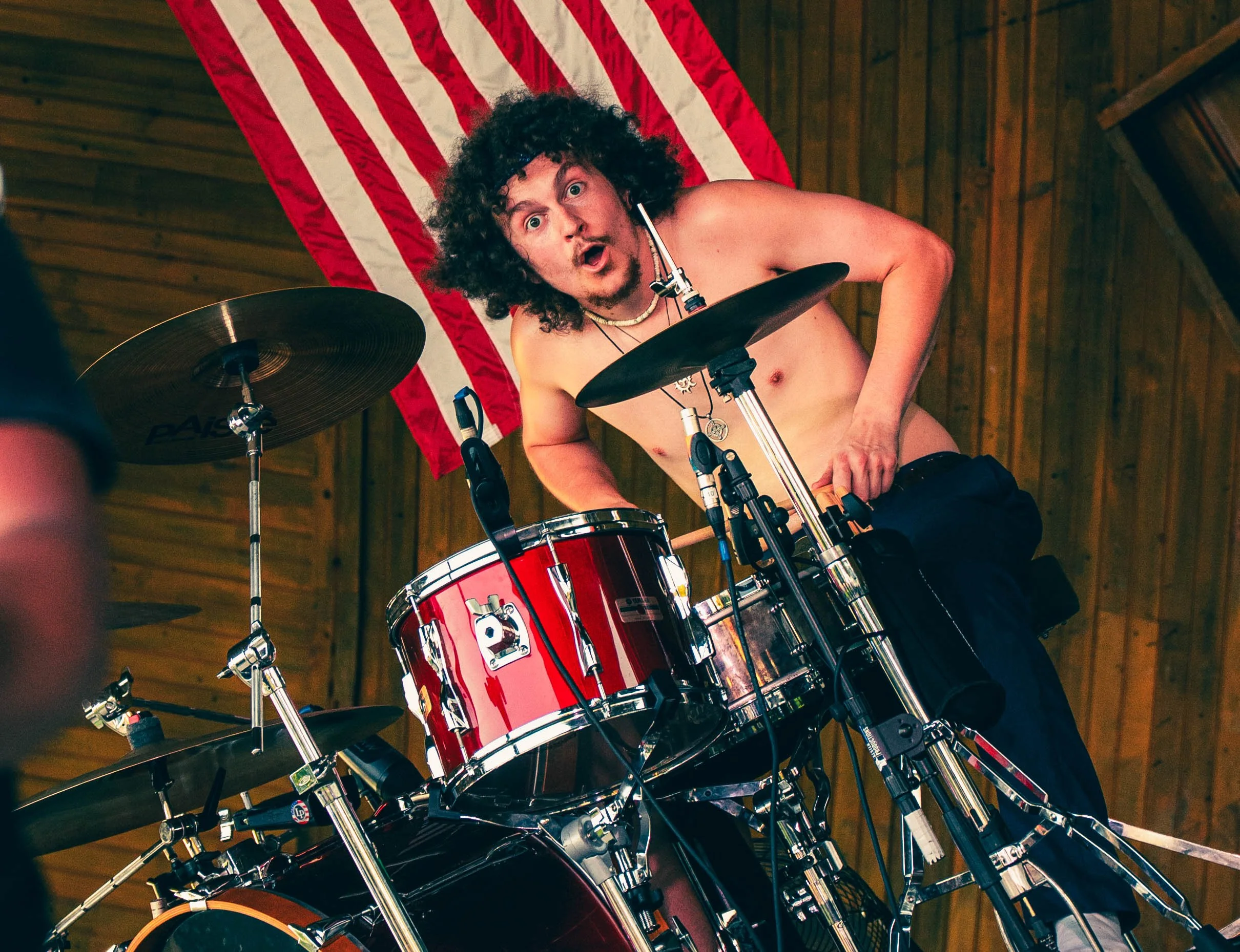 A shirtless man with curly hair and a surprised expression playing a red drum kit with a wooden wall and an American flag in the background.