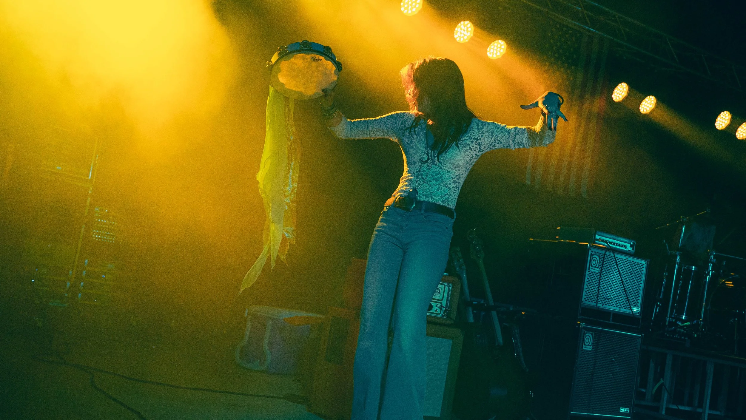 A woman performing on stage, holding a tambourine in one hand and a microphone in the other, with colorful stage lights in the background.