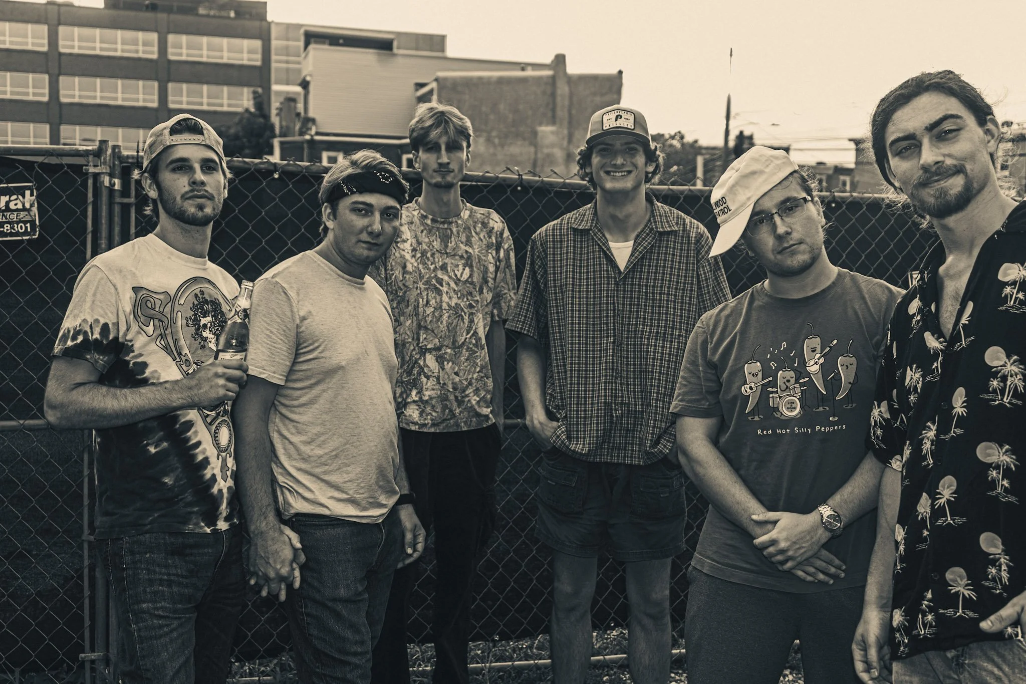 A black-and-white photo of seven young men standing outdoors in front of a chain-link fence, with urban buildings in the background. They are dressed casually, some wearing hats and sunglasses, with relaxed and friendly expressions.