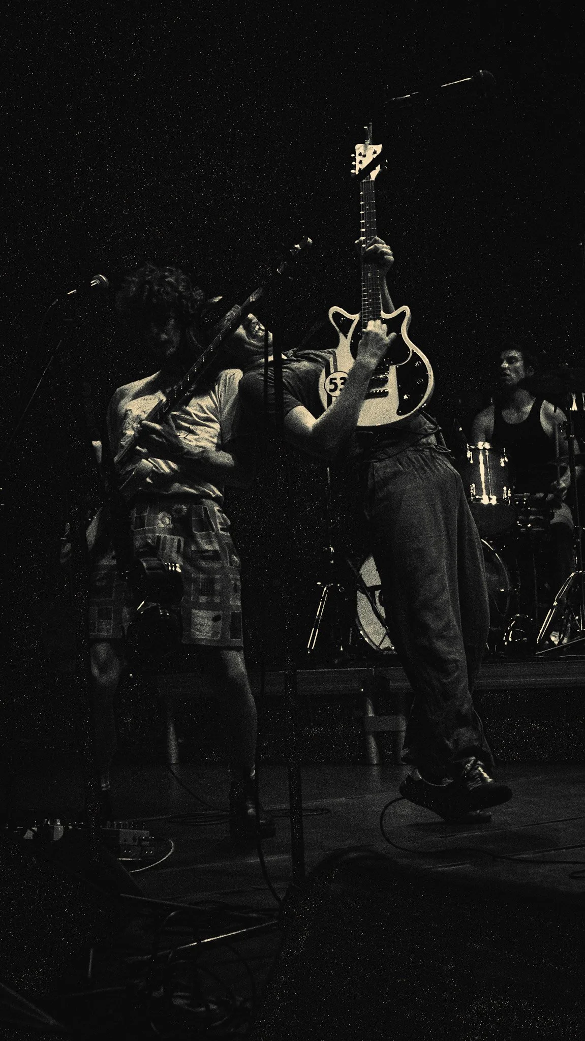 Two musicians performing on stage, one playing a guitar and the other holding a different musical instrument, with a drum set in the background; the scene is in black and white.