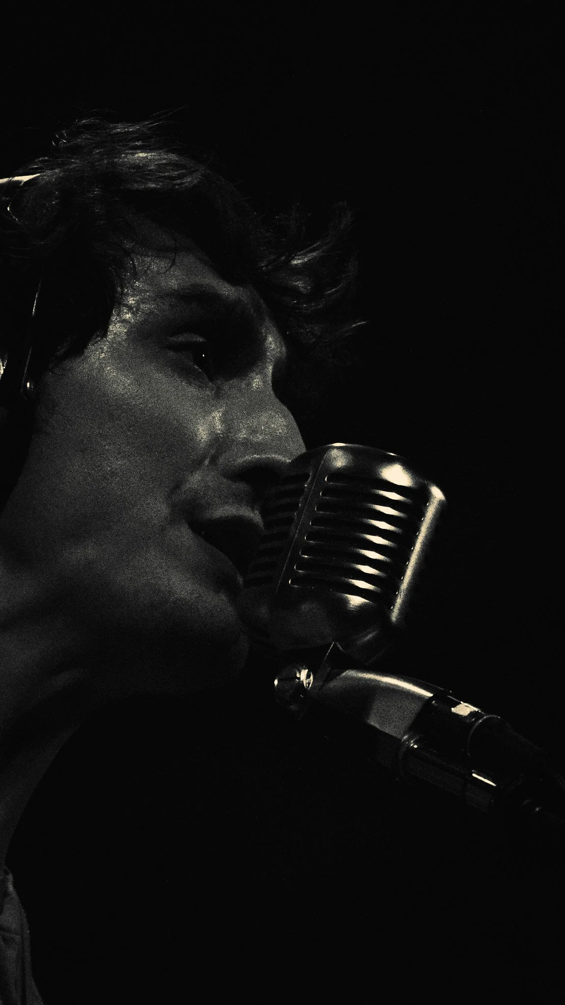 A woman with dark, wavy hair singing into a vintage microphone against a dark background.