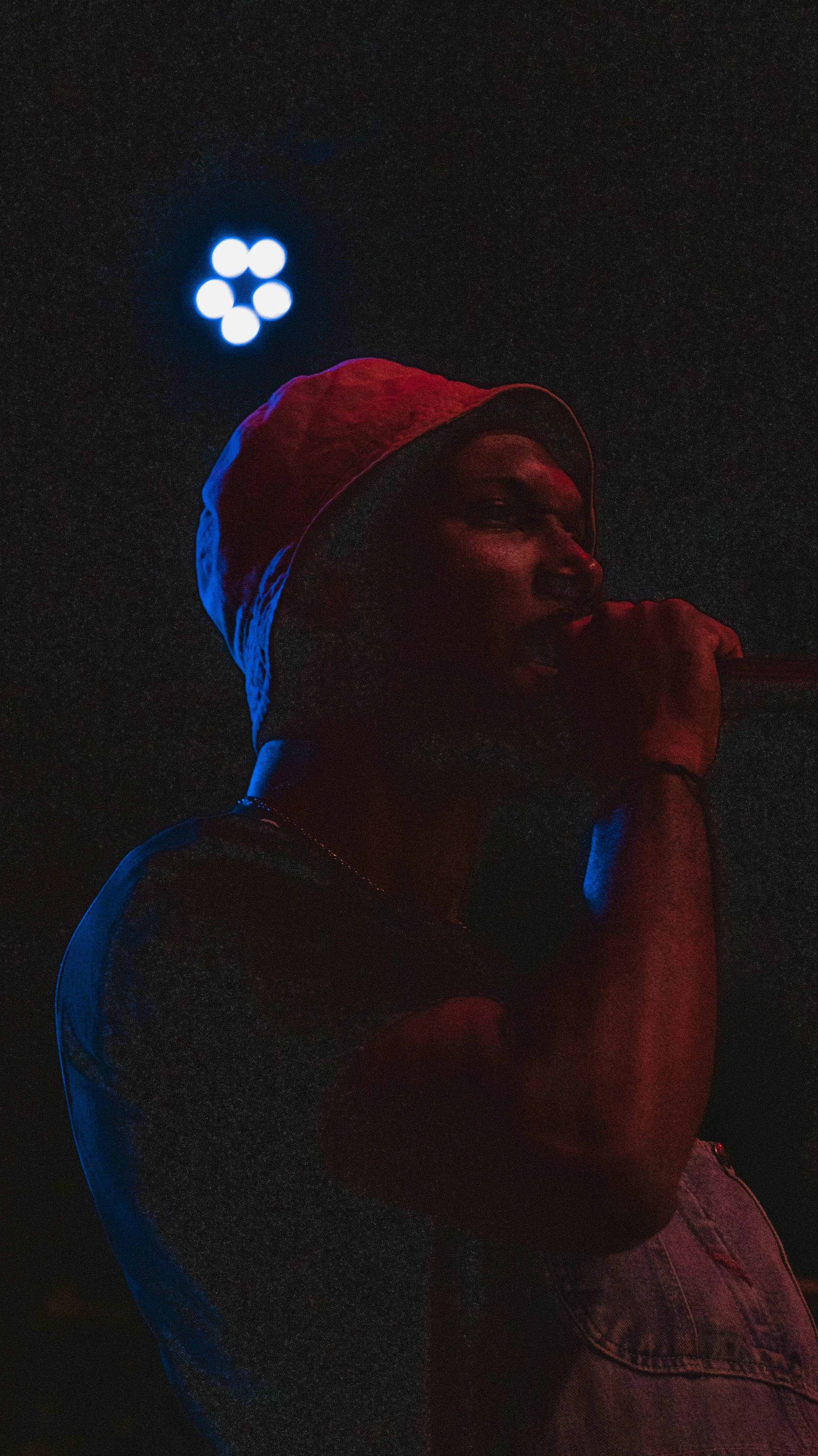 A person wearing a red hat and sleeveless shirt, holding a microphone, standing in a dark setting with blue and red lighting.