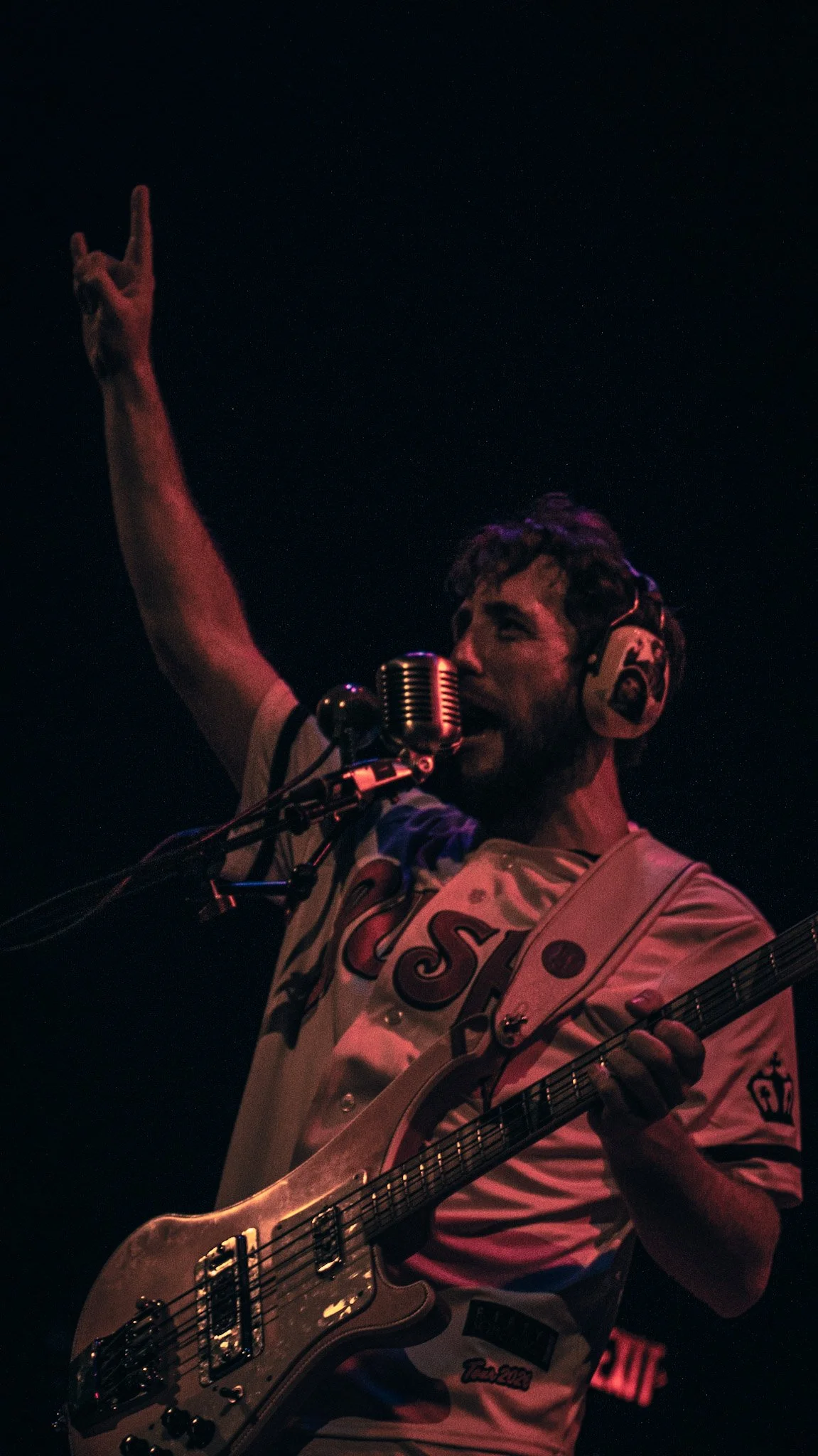 A young man with dark, curly hair and facial hair singing into a microphone and playing an electric guitar on stage. He is wearing a white sports jersey with red and blue accents and a Game of Thrones patch.