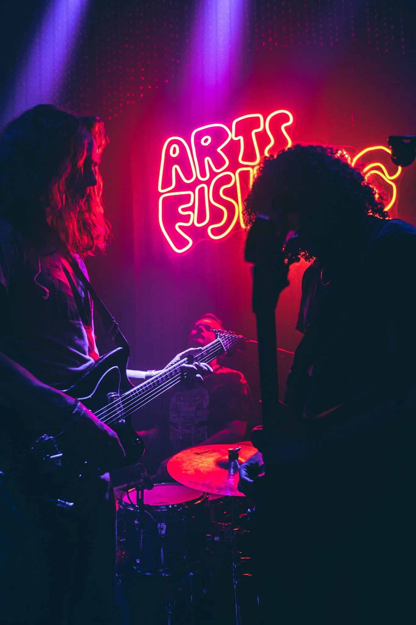 People playing musical instruments in a dimly lit neon-lit art studio, with a sign that reads 'ARTS FEES' in bright neon lights.