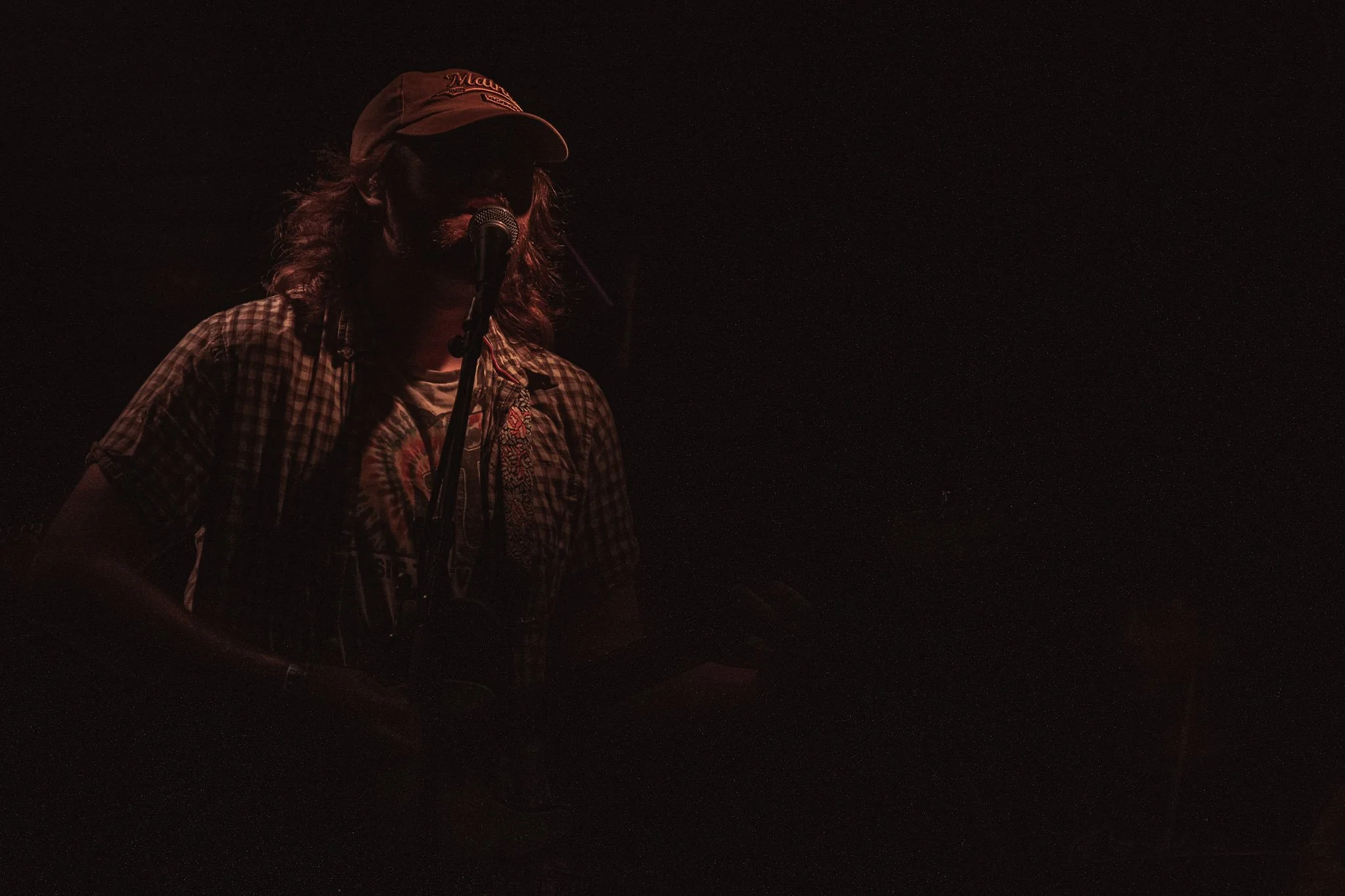 A person wearing a red cap and a plaid shirt is standing in front of a microphone on a dark stage, with their face mostly obscured by shadows.