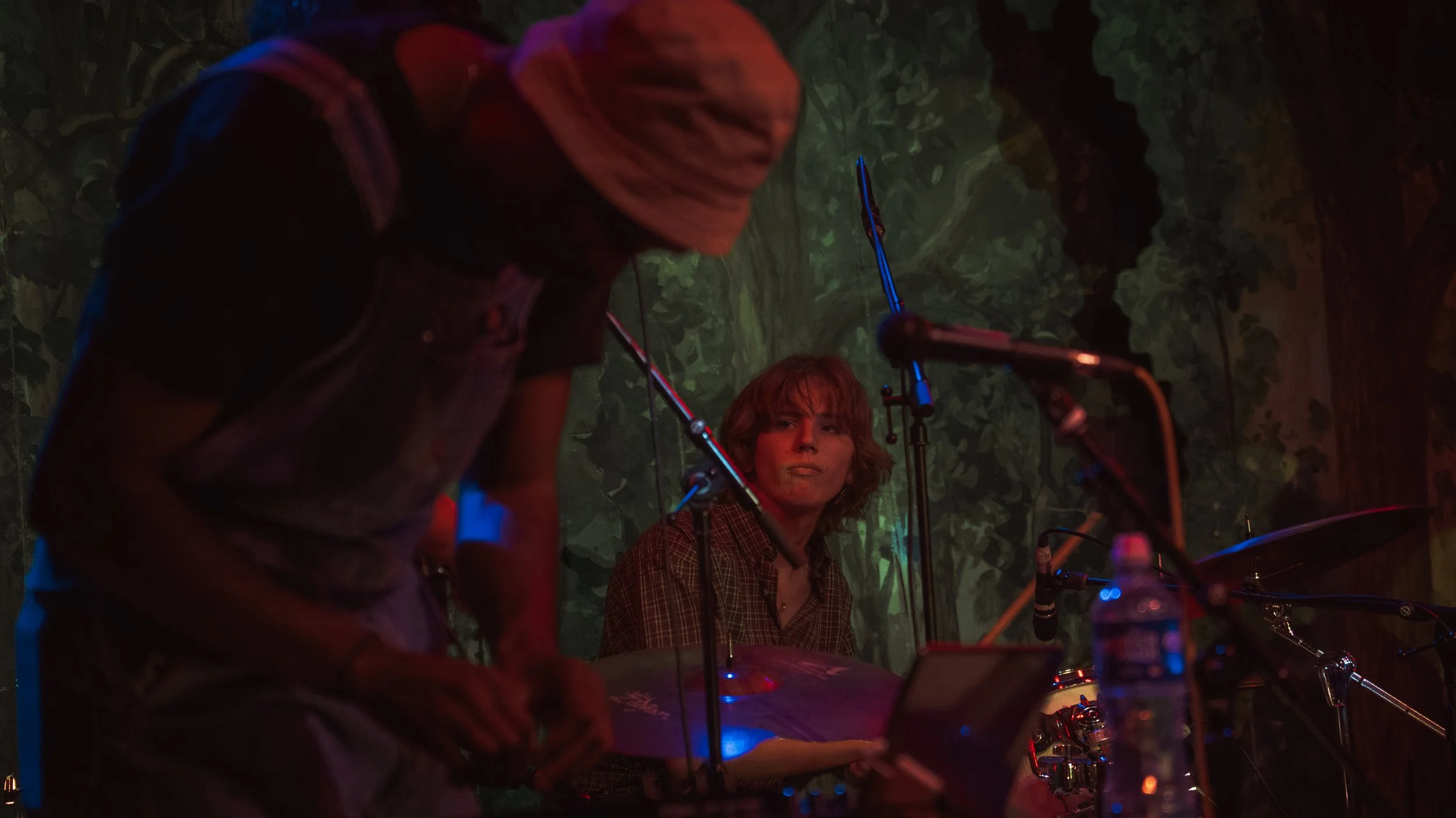 Two musicians performing at a concert, one playing the drums and the other sitting behind a keyboard, surrounded by dark, moody lighting and a backdrop of green foliage.
