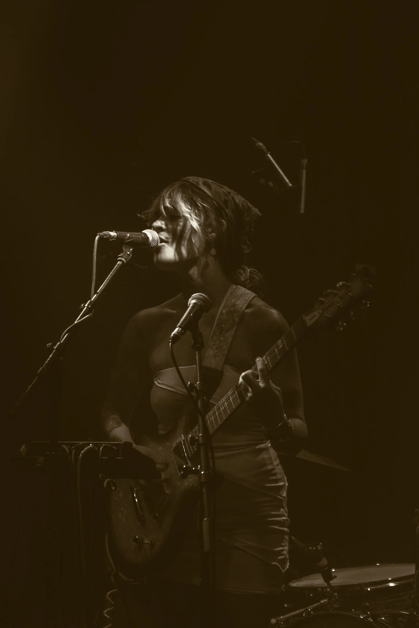 A female musician with wavy hair singing into a microphone while playing an electric guitar on a dark stage.