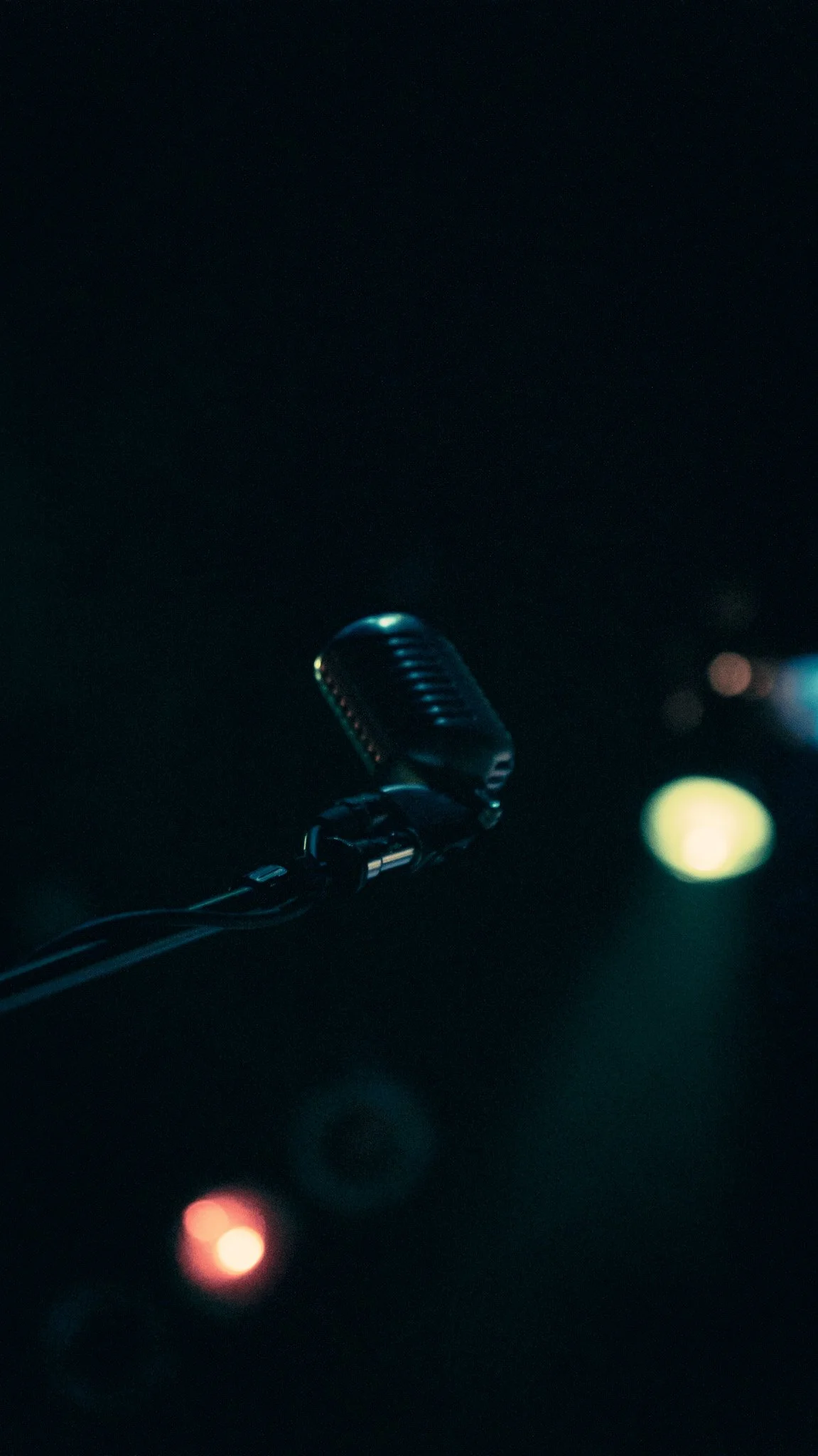 A close-up of a vintage-style microphone illuminated in a dark setting with blurred colorful lights in the background.