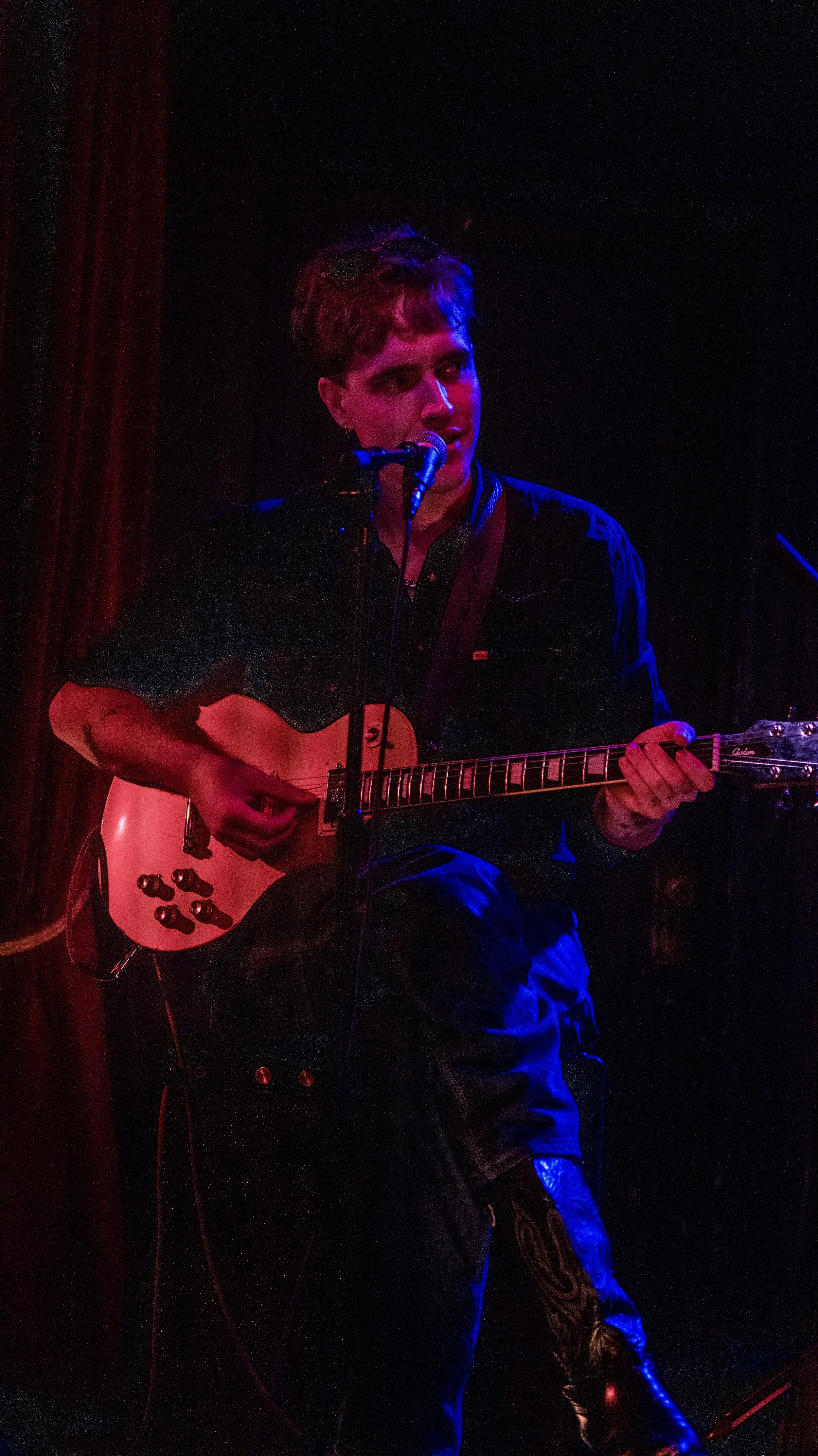 A person with short hair is playing an electric guitar and singing into a microphone on stage. The stage is dimly lit with red and blue lighting.