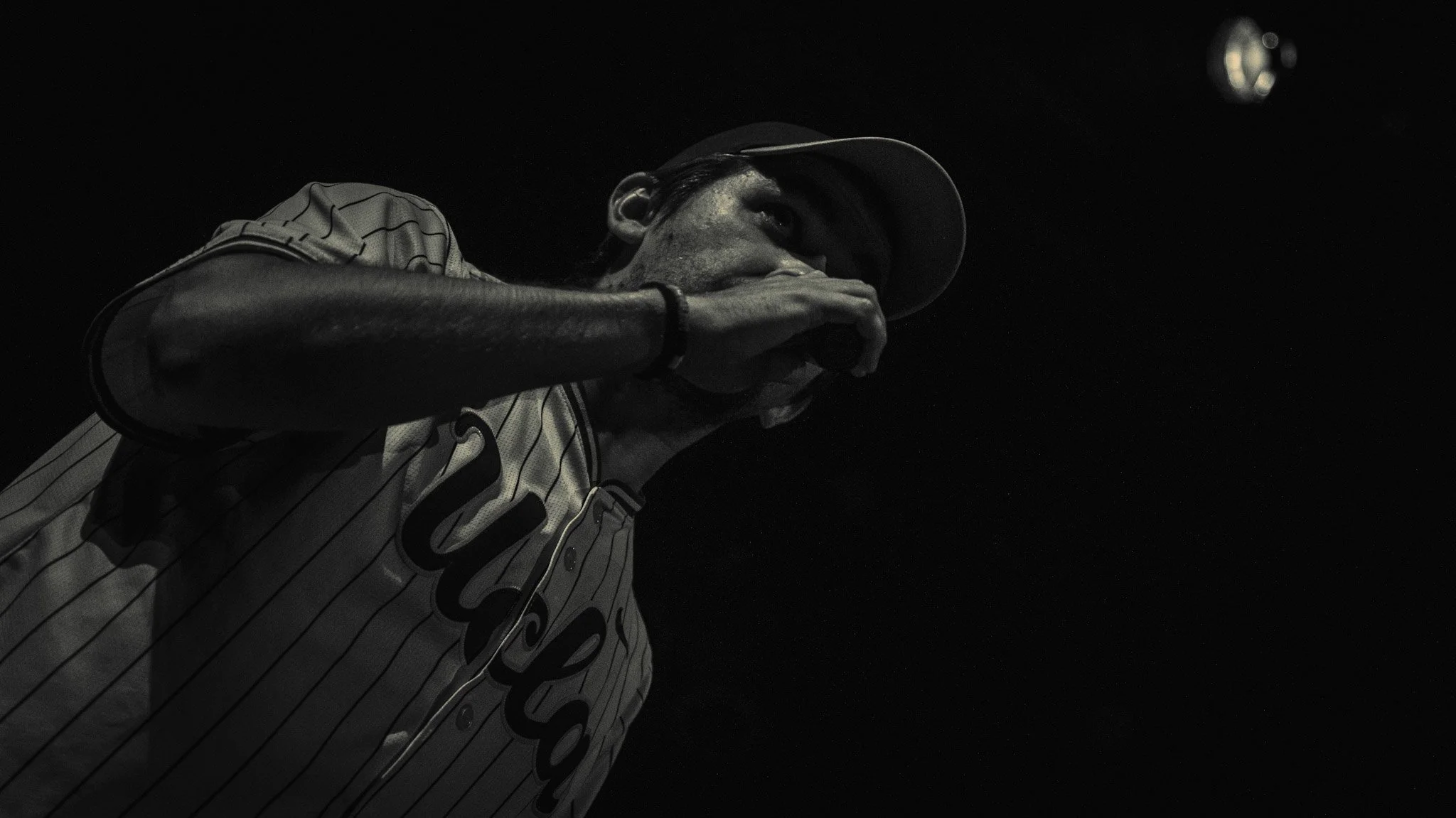 A person wearing a baseball jersey and cap, with earrings, covering part of their face with their hand, in a low-light dark setting.